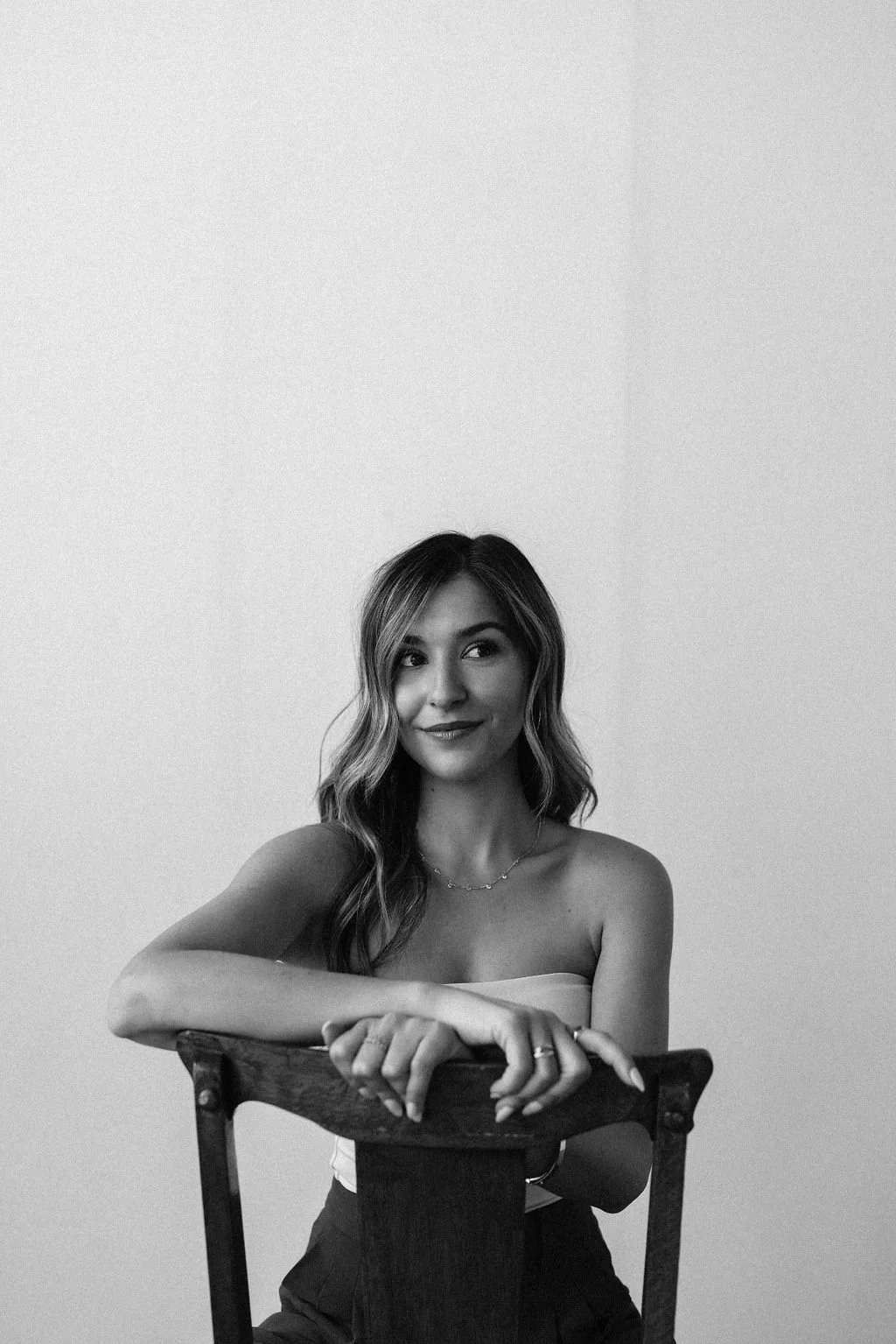 A woman with wavy hair and a strapless top sitting on a wooden chair, looking to her left with a playful smile, in a black-and-white photo.
