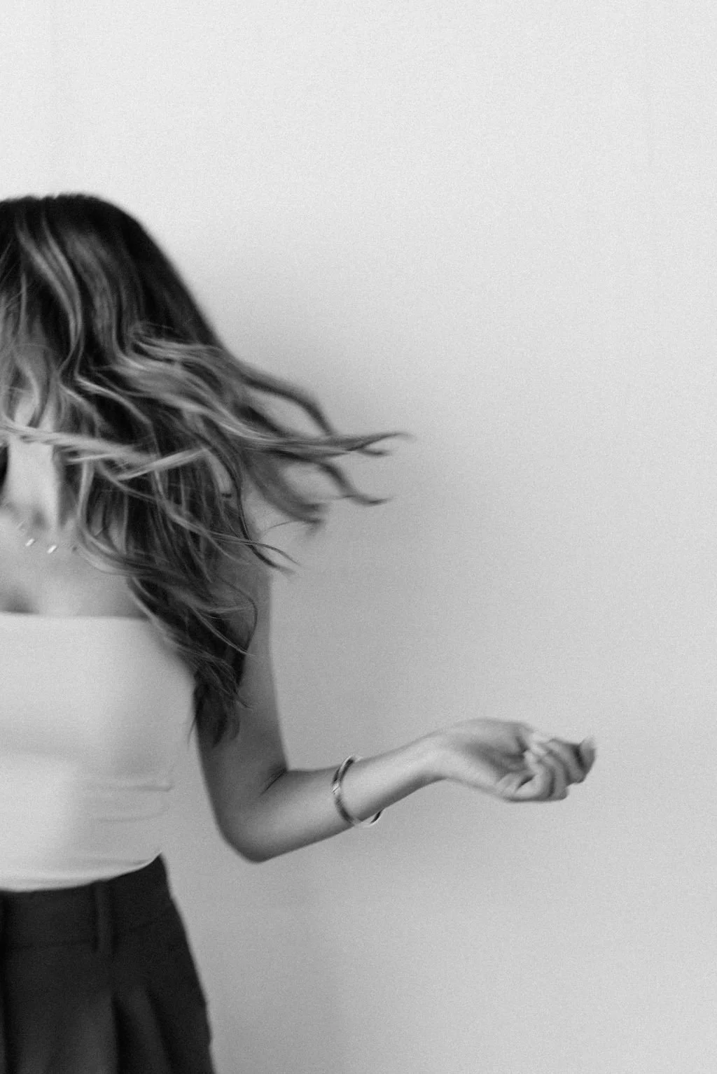 Black and white photo of a woman with long wavy hair, wearing a sleeveless top and a bracelet, moving her head and arm against a plain light-colored background.
