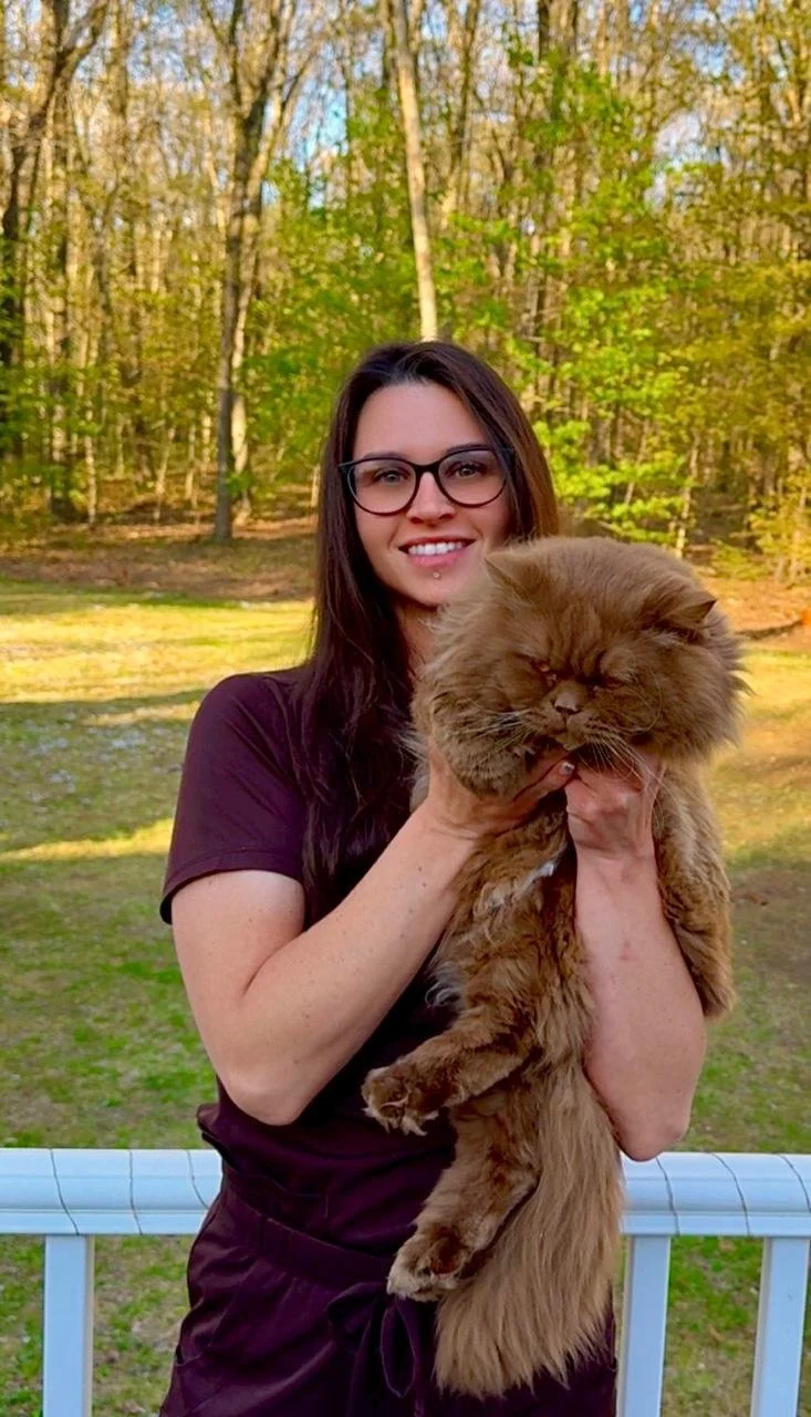 A woman with glasses smiling while holding a fluffy brown cat outdoors during daytime.