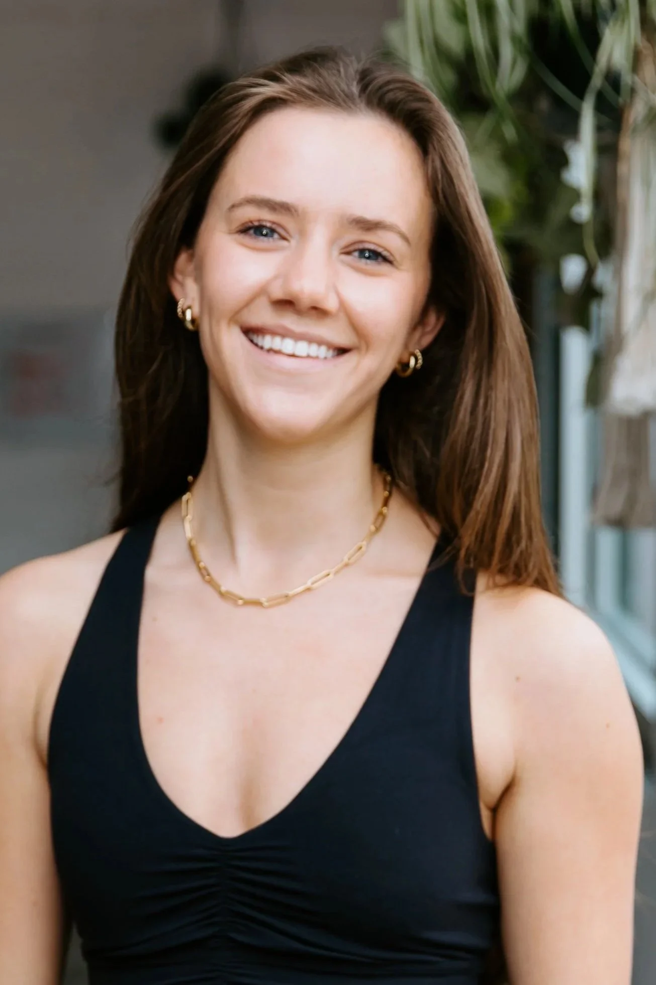 A woman with long brown hair smiles for the camera. She wears a black v-neck tanktop, a gold chain necklace, and small hoop earrings.