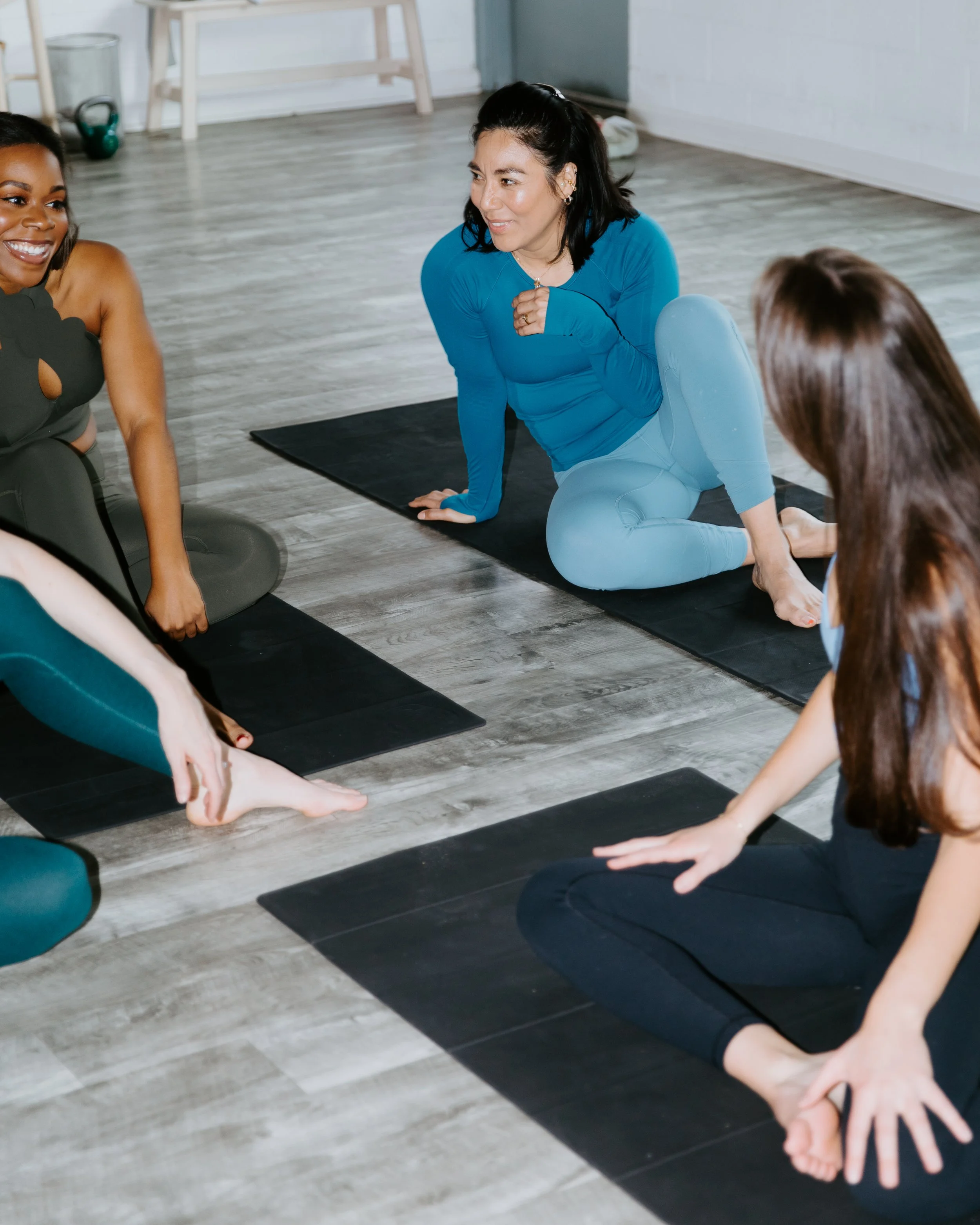 A racially diverse group of women sit on yoga mats, chatting with one another.