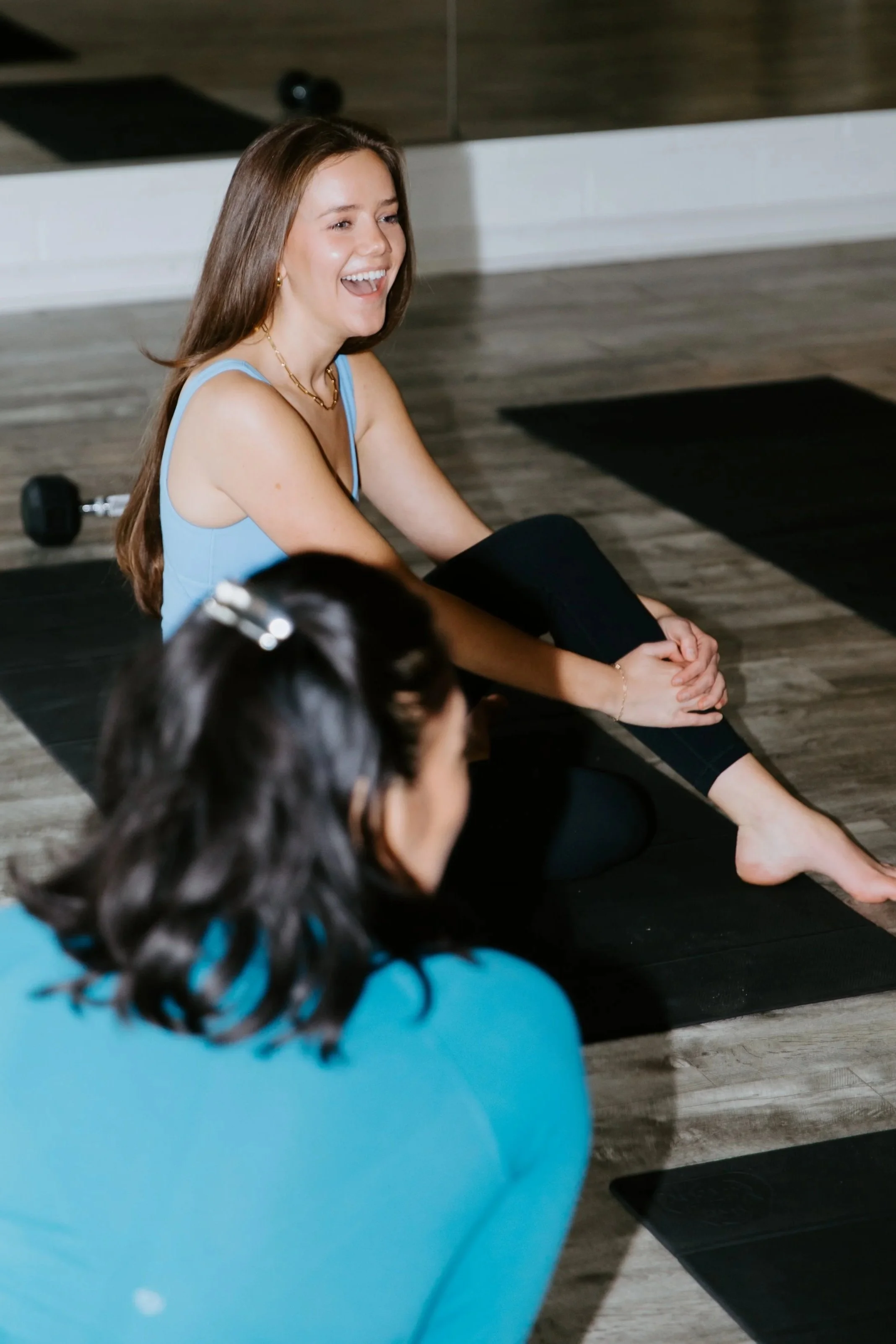 Two women sit on yoga mats, chatting with one another. One is smiling.