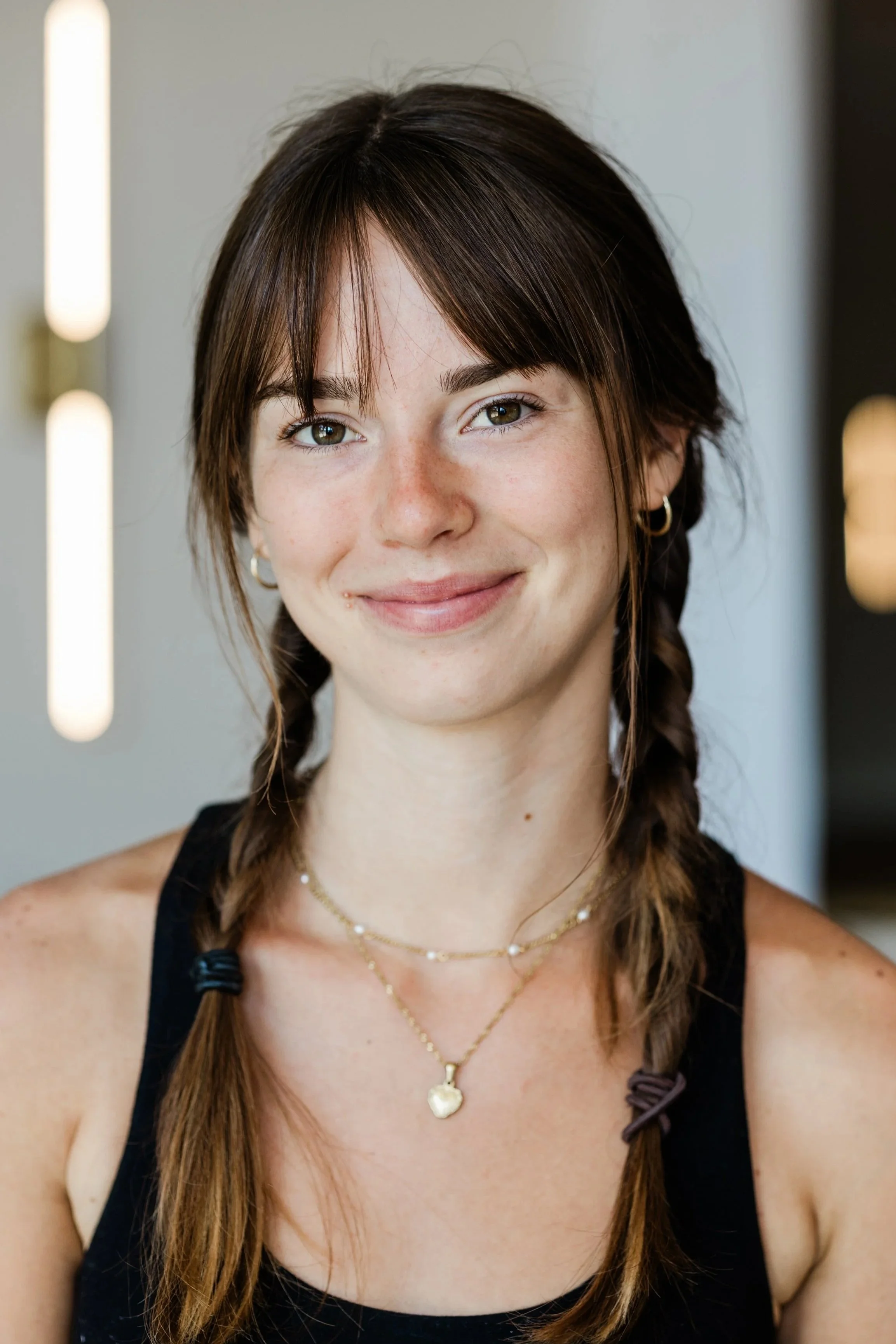A brunette woman with bangs and two long side braids smiles for the camera. She wears a black tank top, two layered necklaces and small silver hoop earrings.