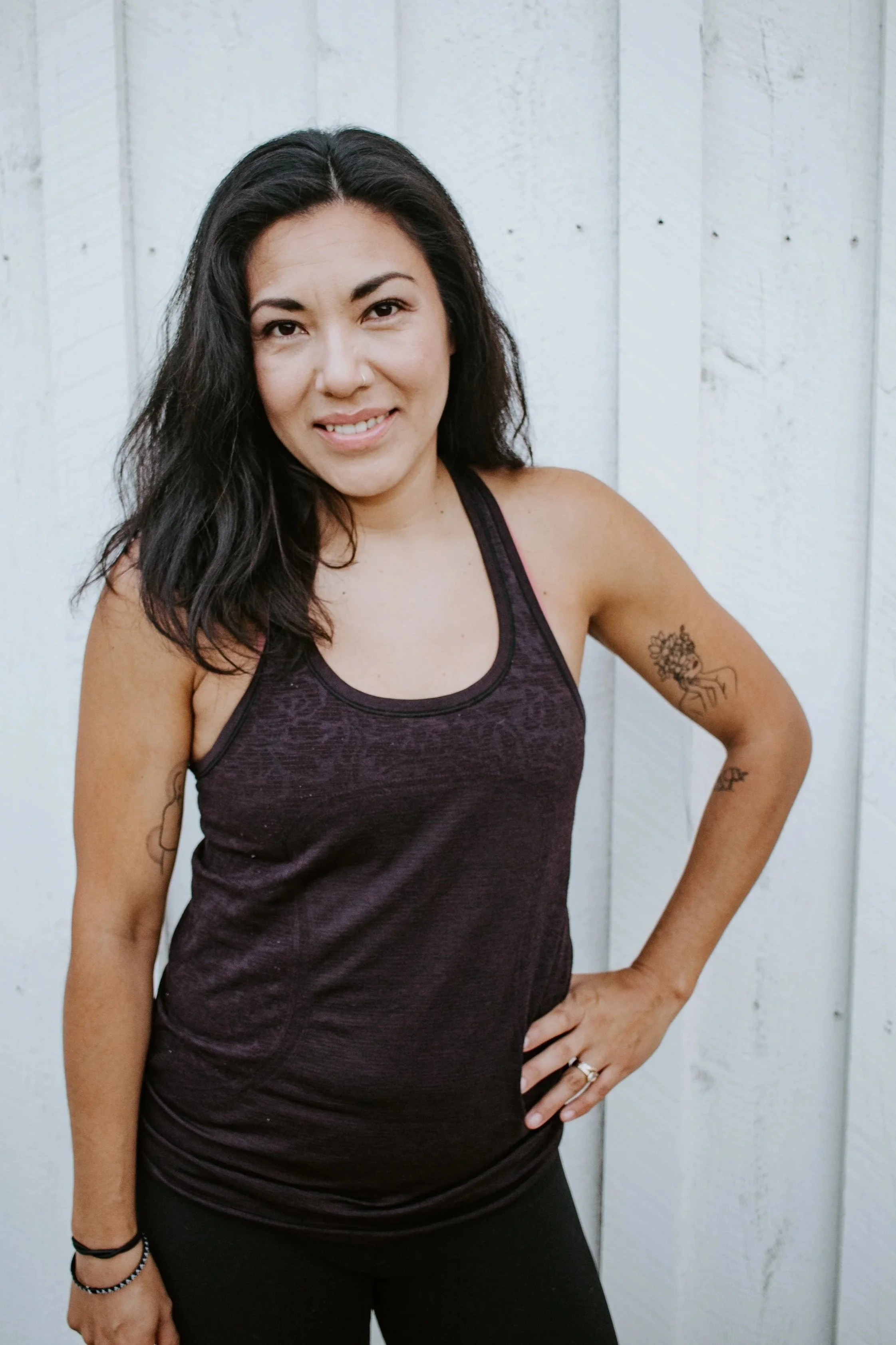 A woman with long dark hair smiles for the camera. One hand is on her hip, and she has a tattoo on her upper arm.
