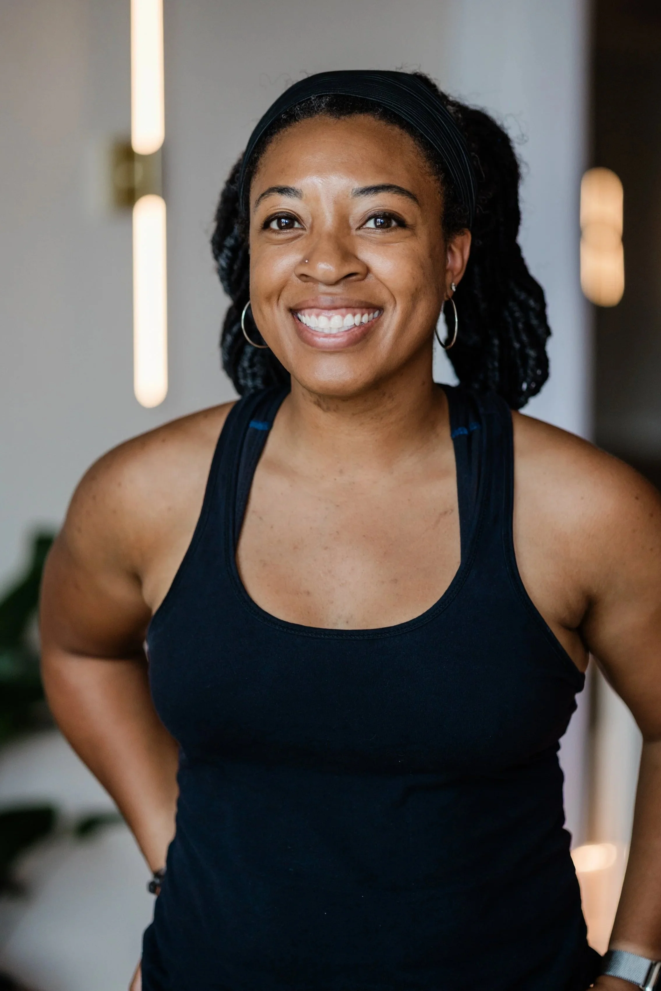 A woman with black, braided, pulled back hair smiles for the camera. She wears a black tank top and small silver hoop earrings,  and holds her hands on her hips. .
