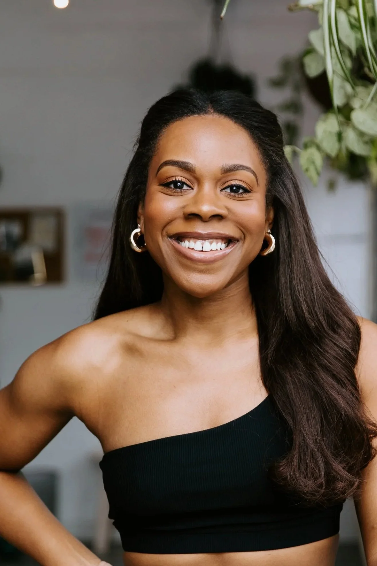 A woman with long black hair worn half up, half down smiles for the camera. She wears a black one-shoulder crop top and medium-sized silver hoop earrings.