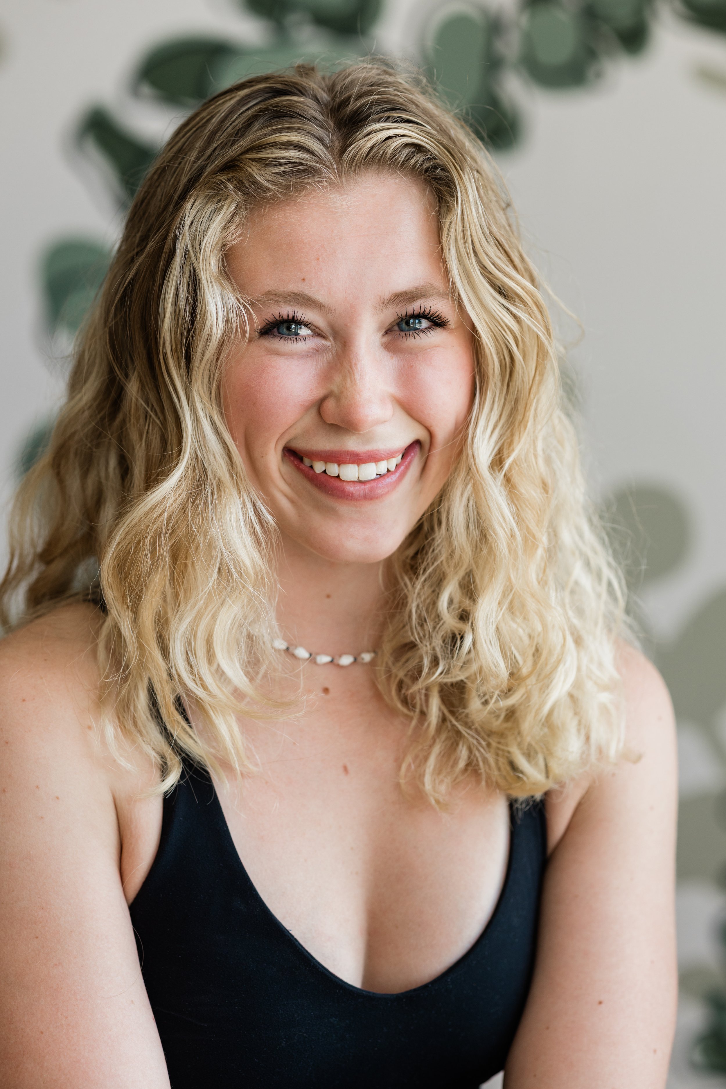 A woman with long, wavy blonde hair smiles for the camera. She wears a black tank top and a white beaded necklace.