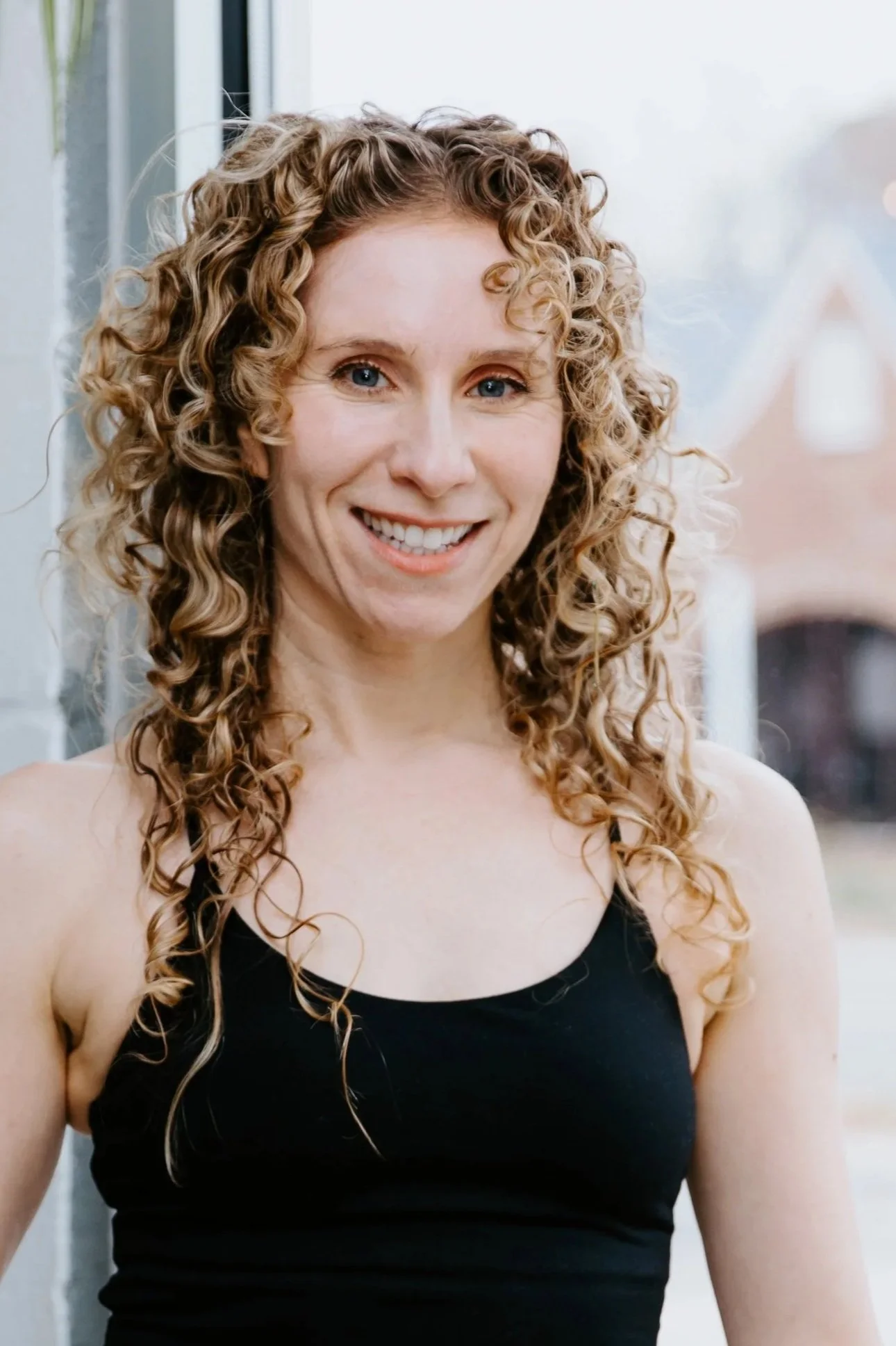A woman with long blonde curly hair worn half up, half down smiles for the camera. She wears a black tank top.