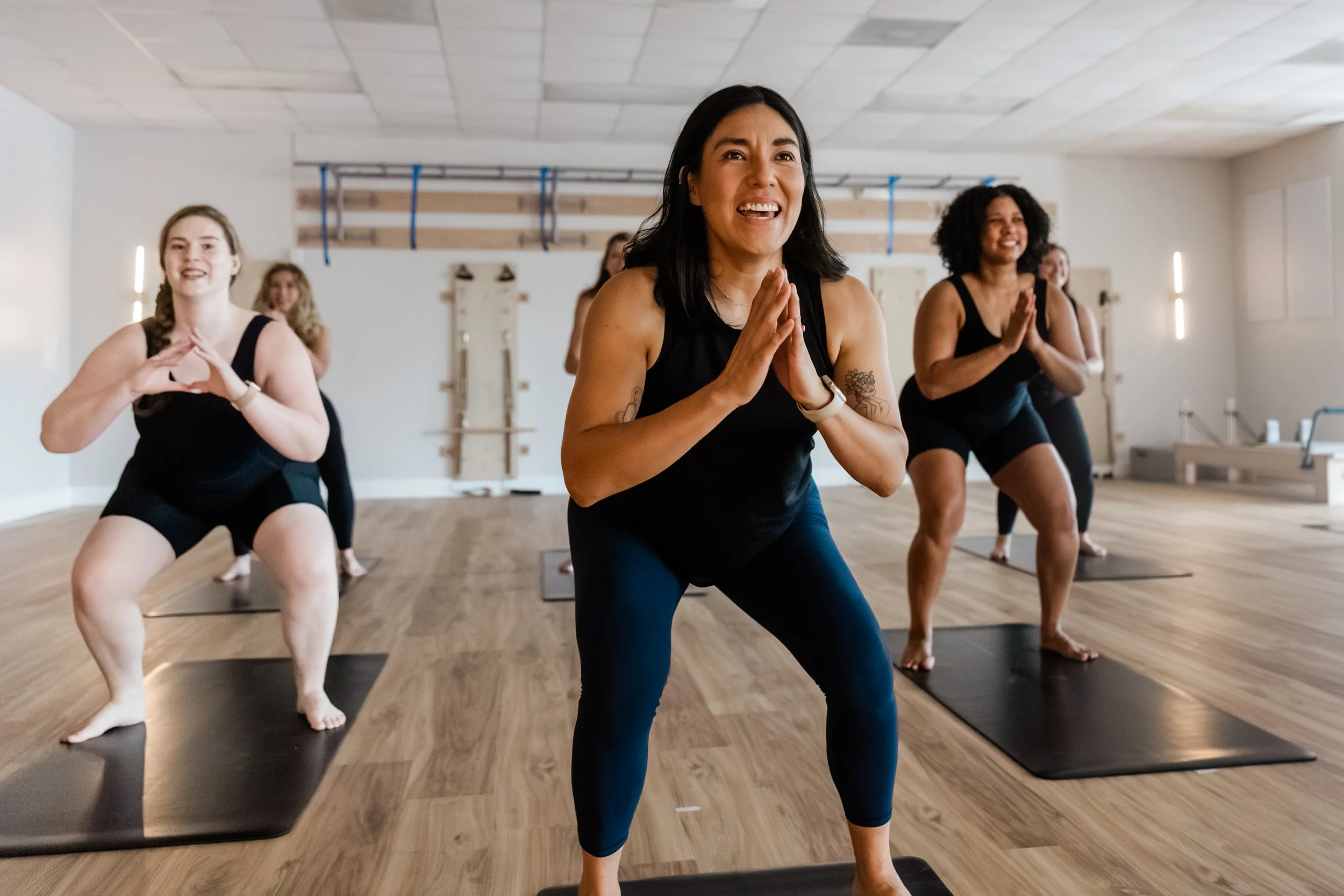 A group of women take a fitness class. They sink into a deep squat and all are smiling.