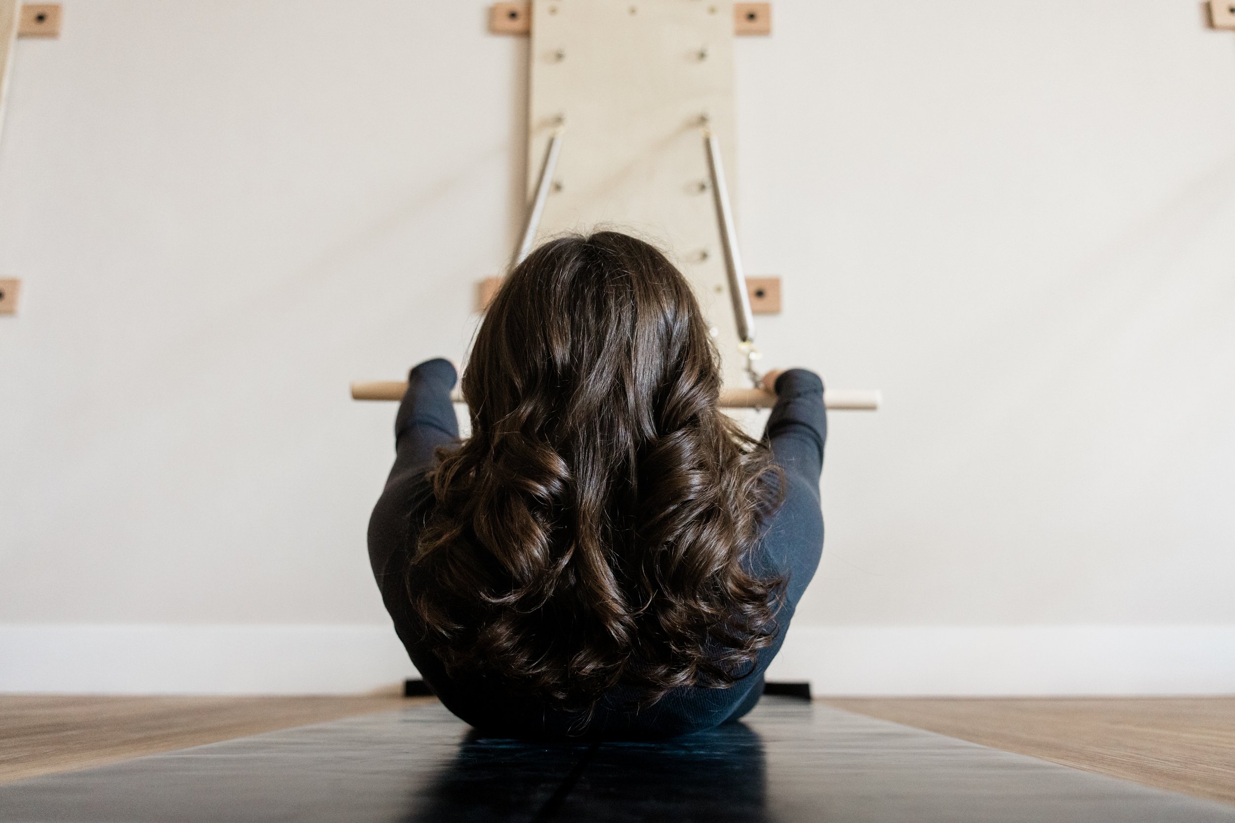A woman with long dark hair is seen from the back. She is rolling up on a yoga mat, hands holding a bar attached to springs.