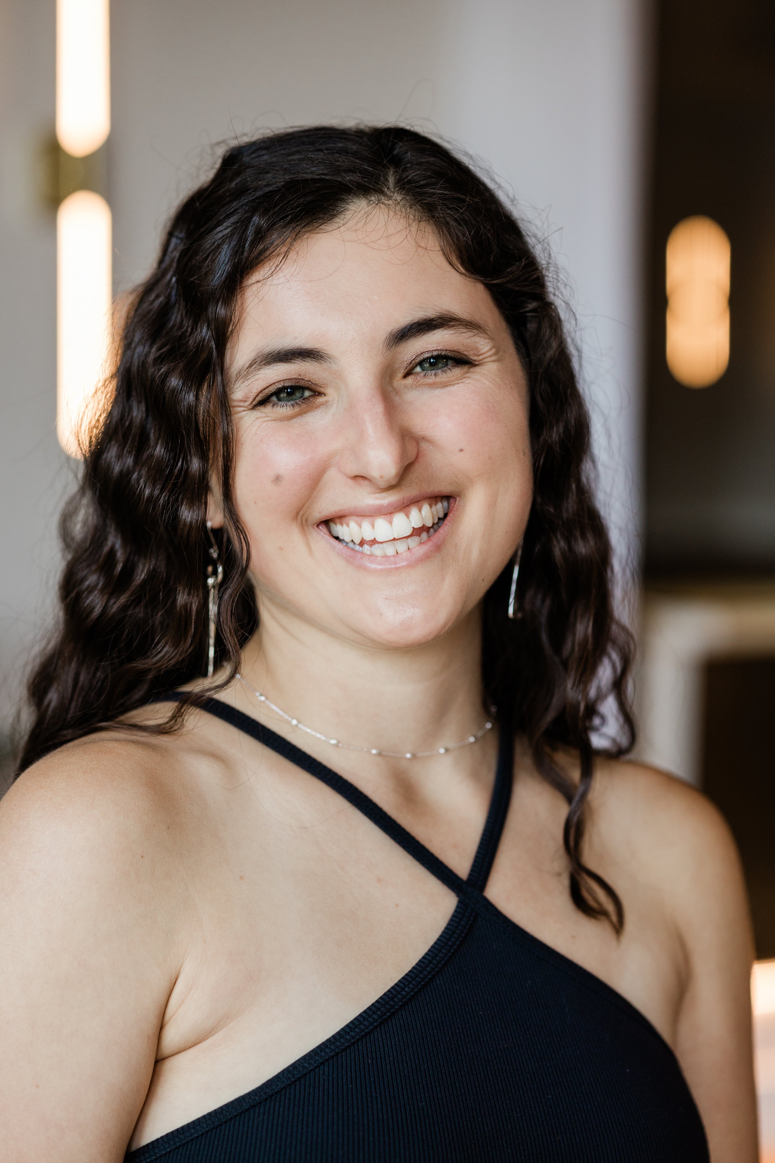 A woman with long, wavy brown hair smiles for the camera. She wears a black tank top, dangly silver earrings, and a white beaded necklace.