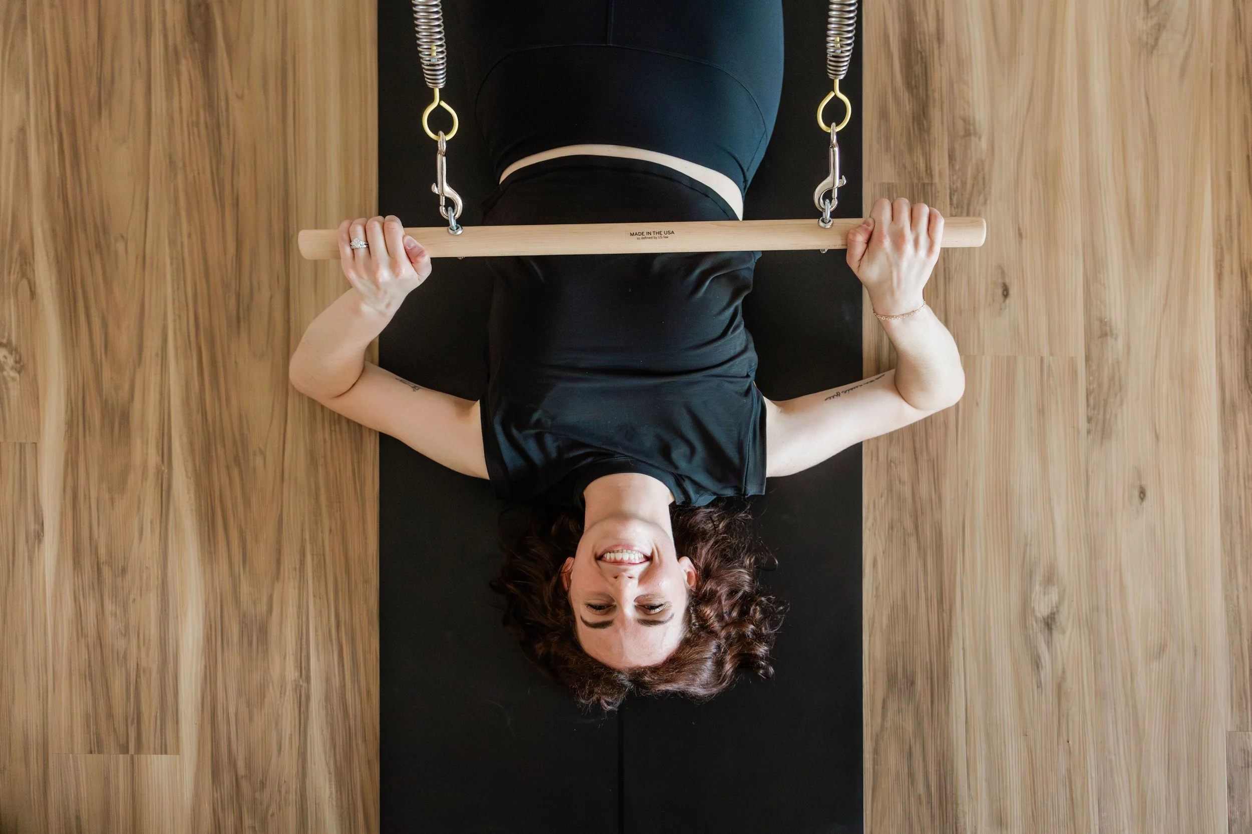 A woman with dark hair lies on a yoga mat, smiling. She is seen from overhead. She holds a bar with springs attached to it.