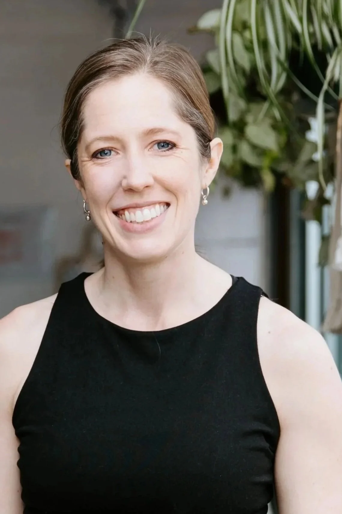 A woman with light brown hair worn pulled back smiles for the camera. She wears a black tank top and small silver hoop earrings.