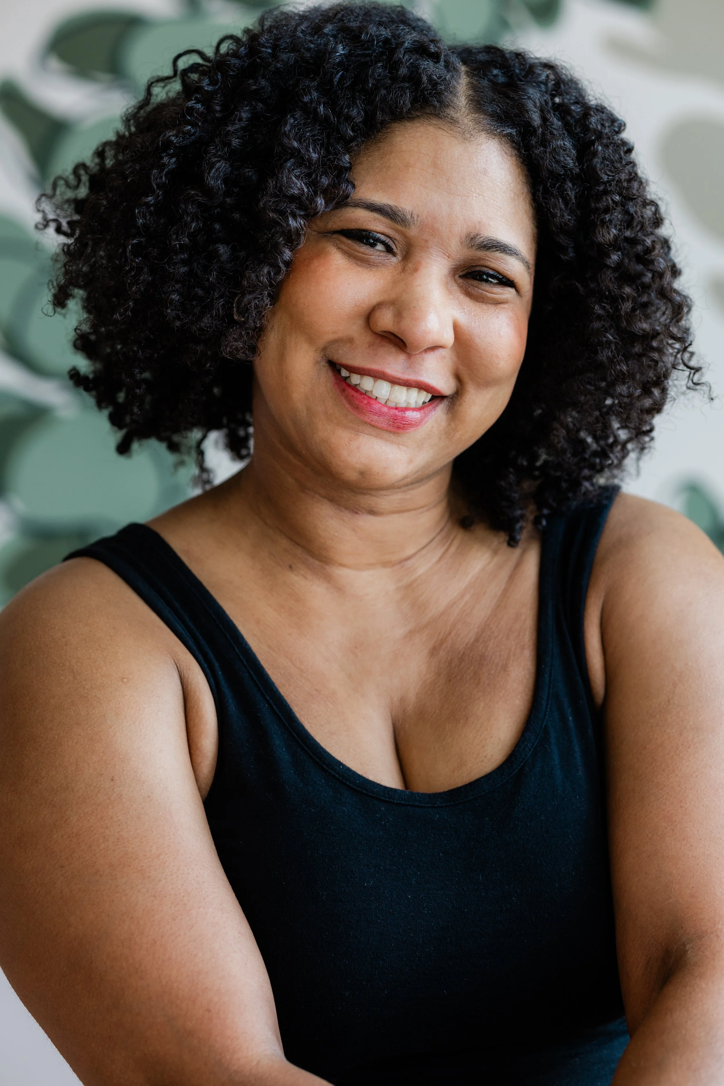 A woman with medium length, black curly hair smiles for the camera. She wears a black tank top.