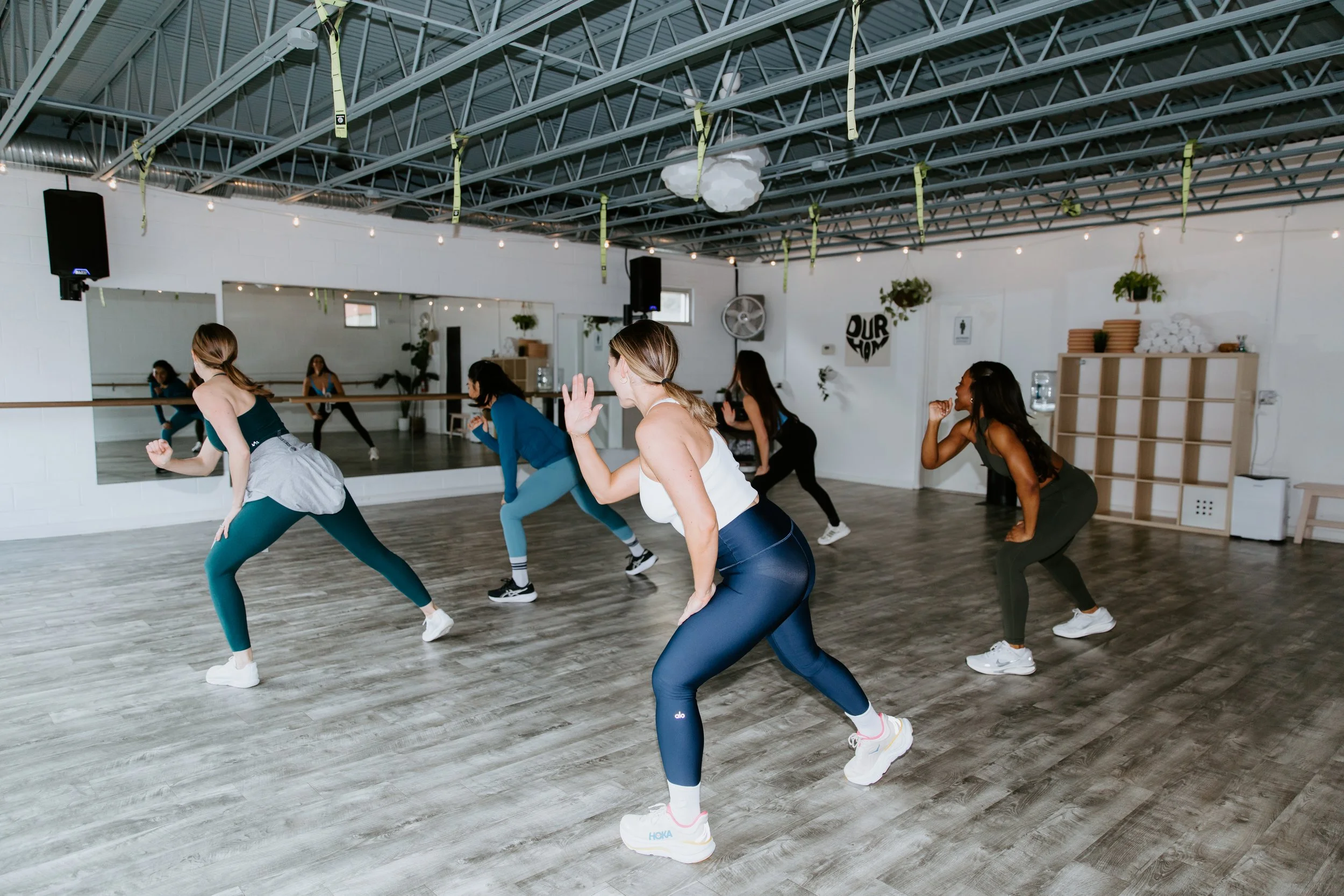 A group of women wearing colorful fitness clothing participate in a Zumba class. They lean to one side and pump the opposite arm. They are in a big open studio.