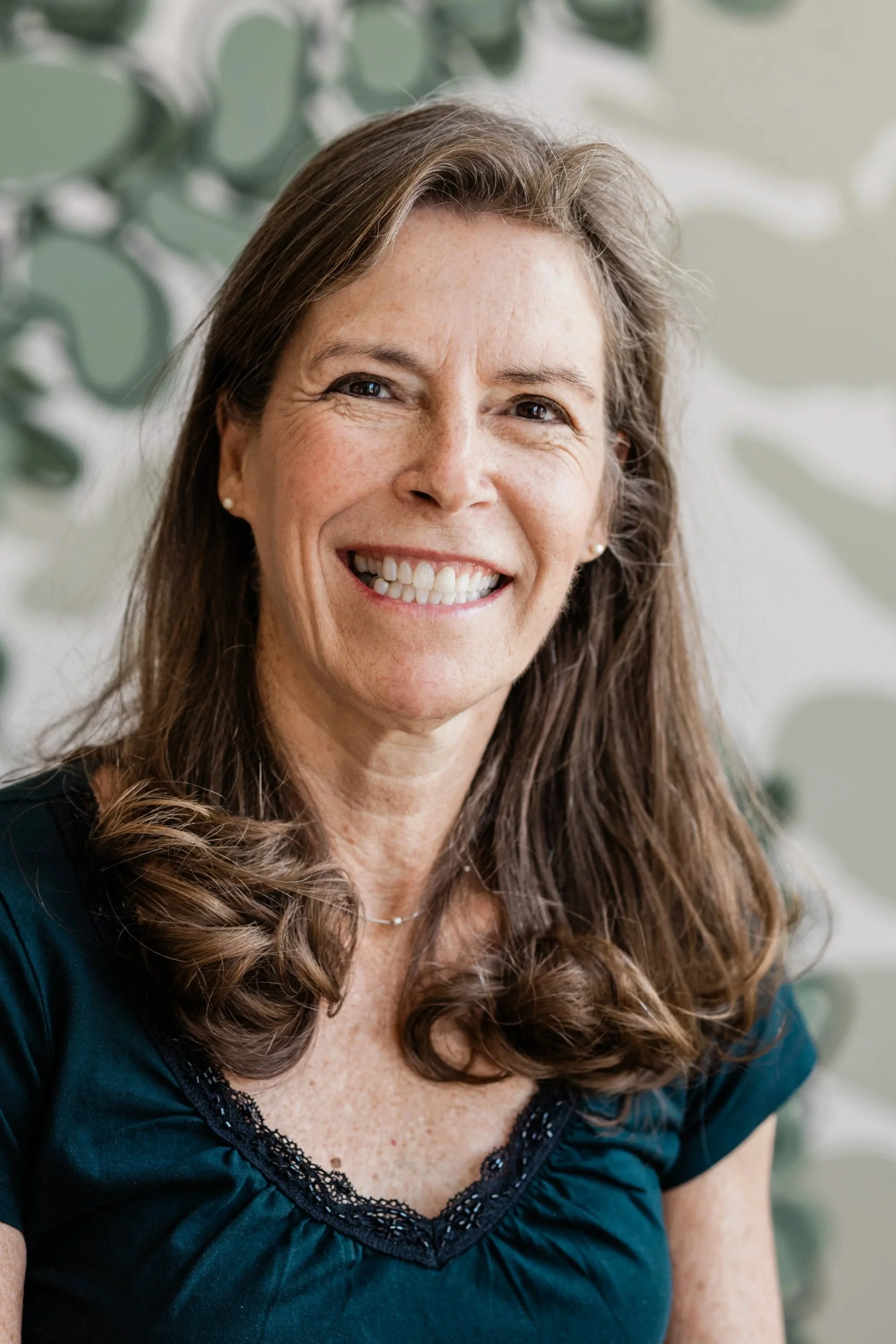 A woman with long, light brown, straight hair smiles for the camera. She wears a black t-shirt with a ruffle and small pearl earrings.