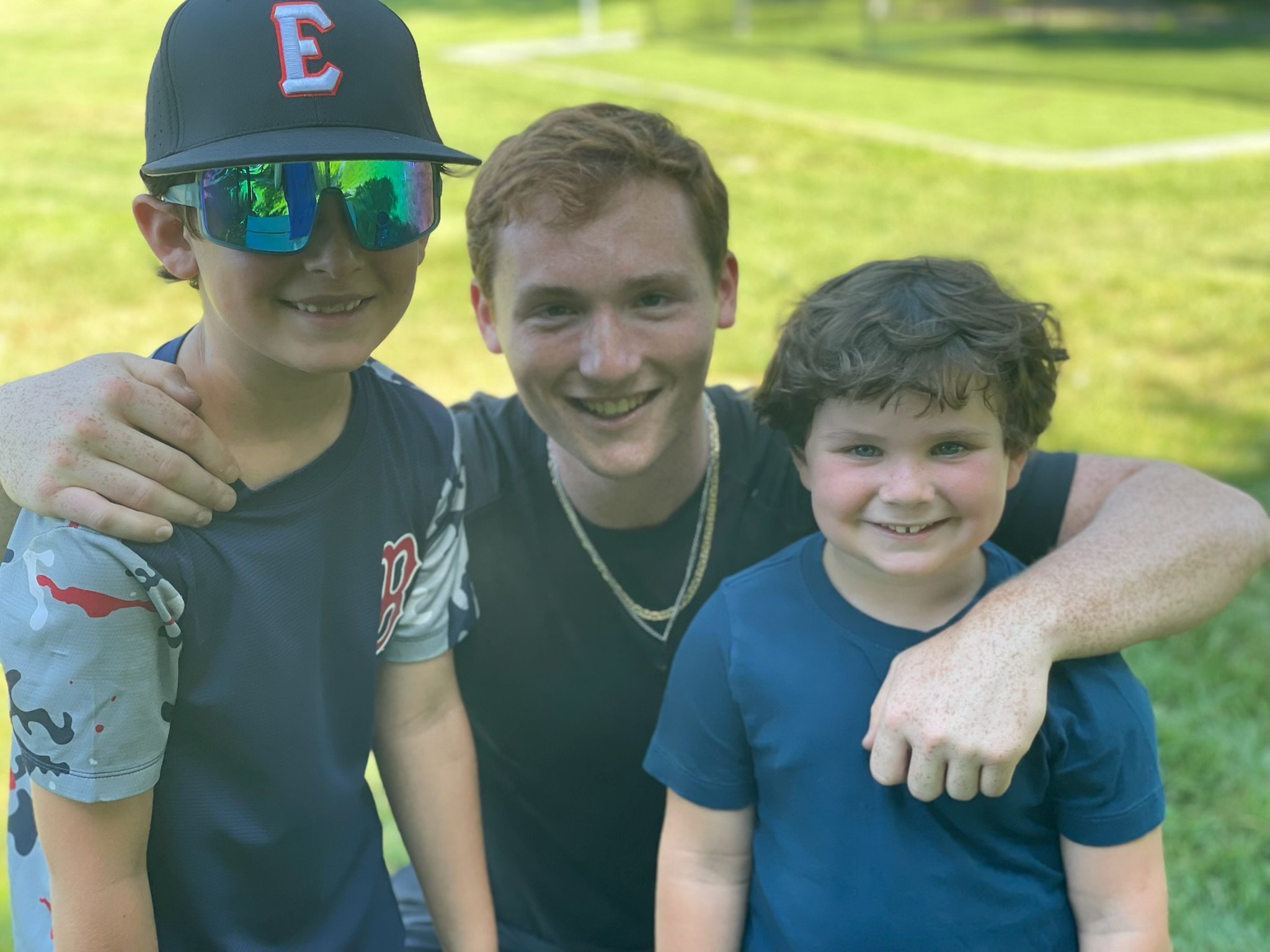 Three boys and a man smiling outdoors, with one boy wearing sunglasses and a baseball cap. The man is in the middle, arms around two boys, all smiling.