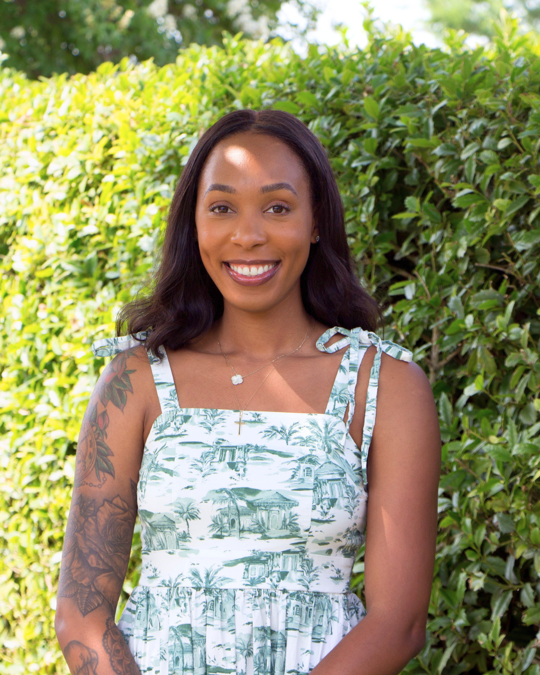 A young woman with dark hair, smiling outdoors in front of green shrubbery, wearing a white dress with a scenic print and tied shoulder straps, and gold jewelry.