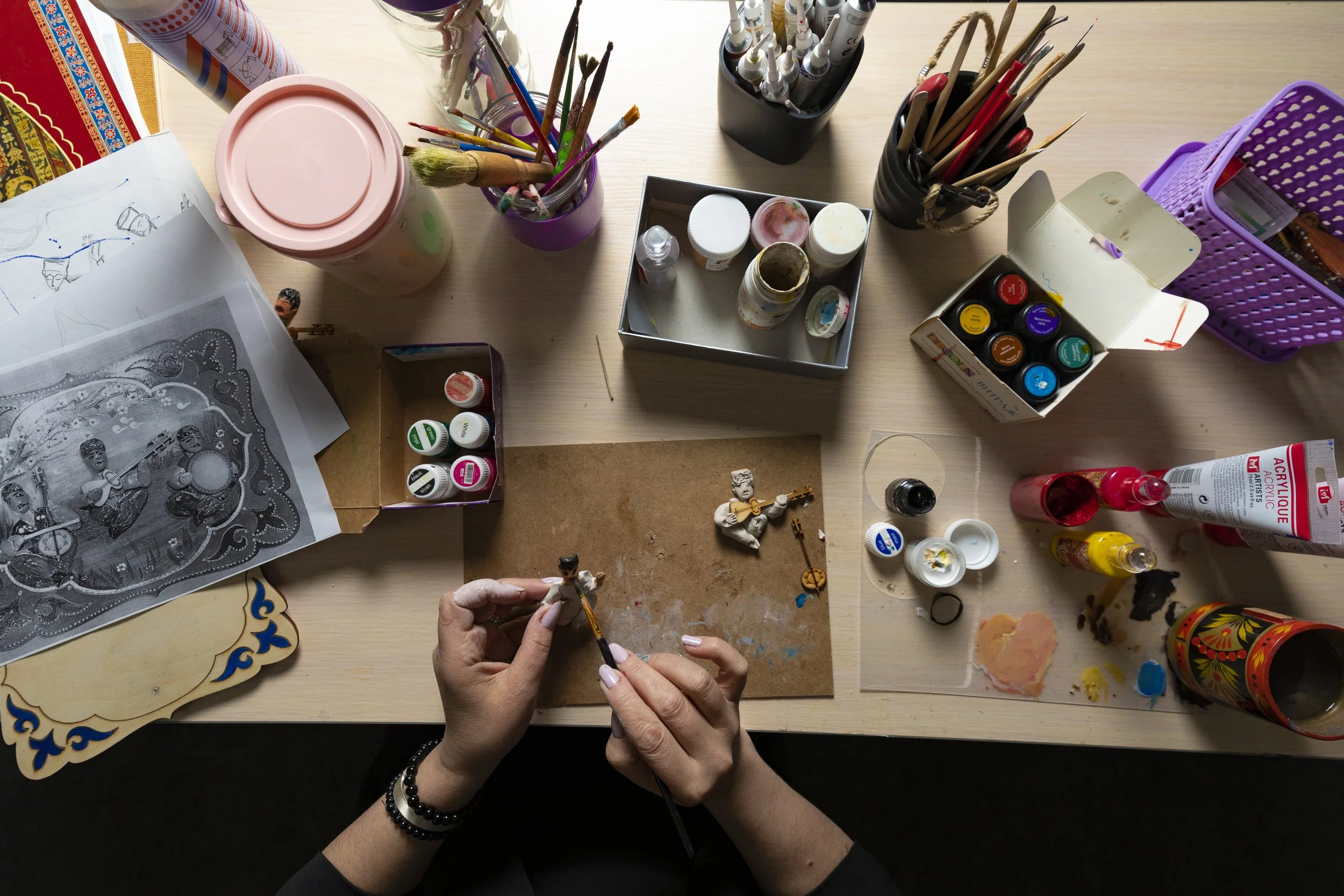 A person working on miniature figurines with paint and brushes on a cluttered desk, surrounded by paint bottles, art supplies, and drawings.