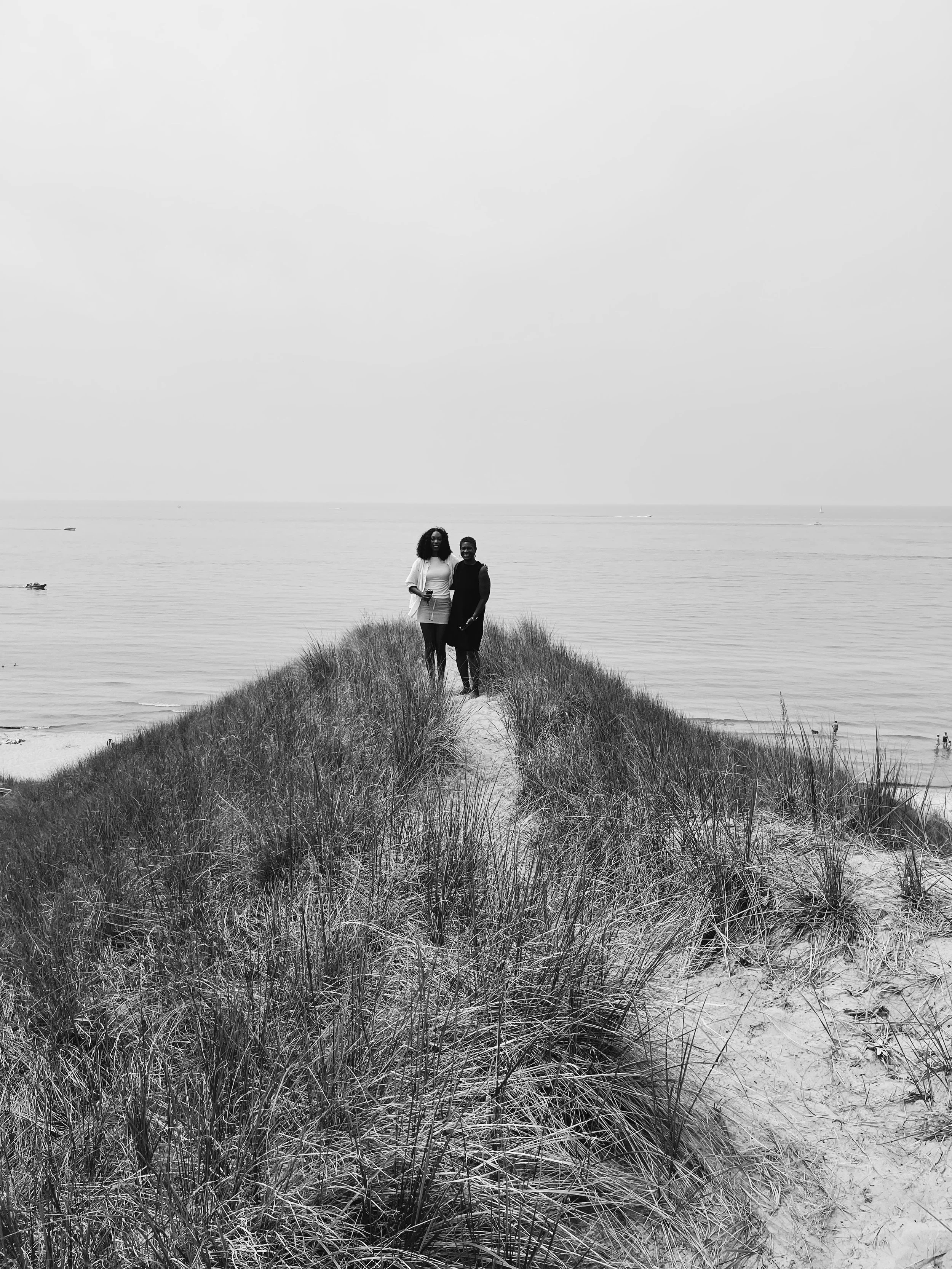 Two women standing together on a narrow sand path between beach grass, at a beach with the ocean in the background.