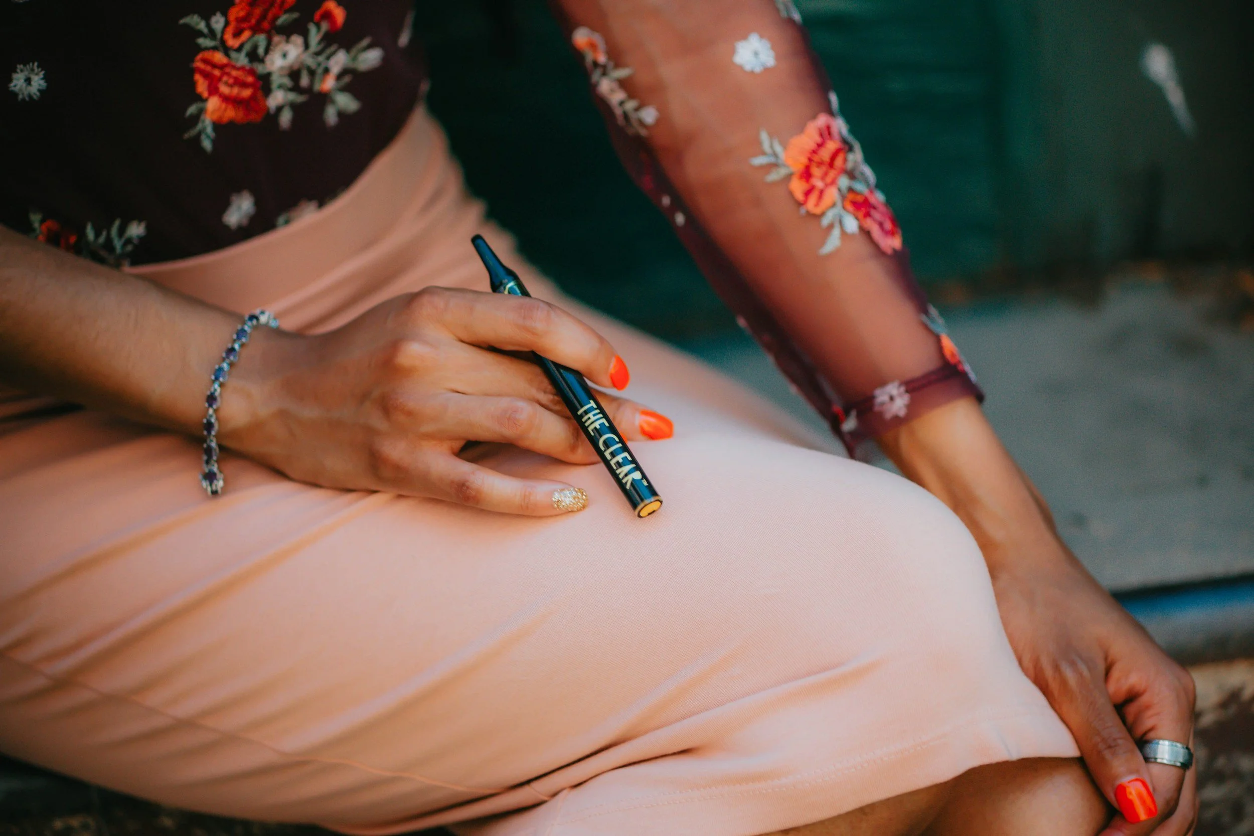 Close-up of a woman writing with a marker on someone's skin. The woman is wearing a floral shirt with orange and white flowers, a bracelet, and has bright orange painted nails. The skin appears to belong to a child wearing peach-colored clothing.