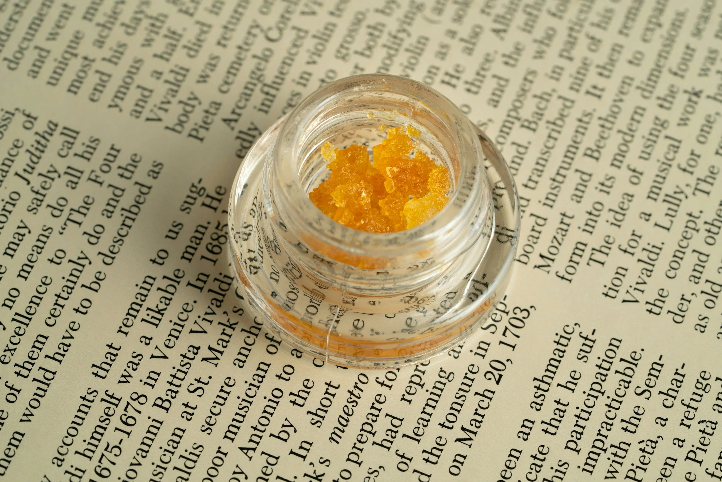 A small glass bottle with yellowish-orange crystals on top, placed on a newspaper.