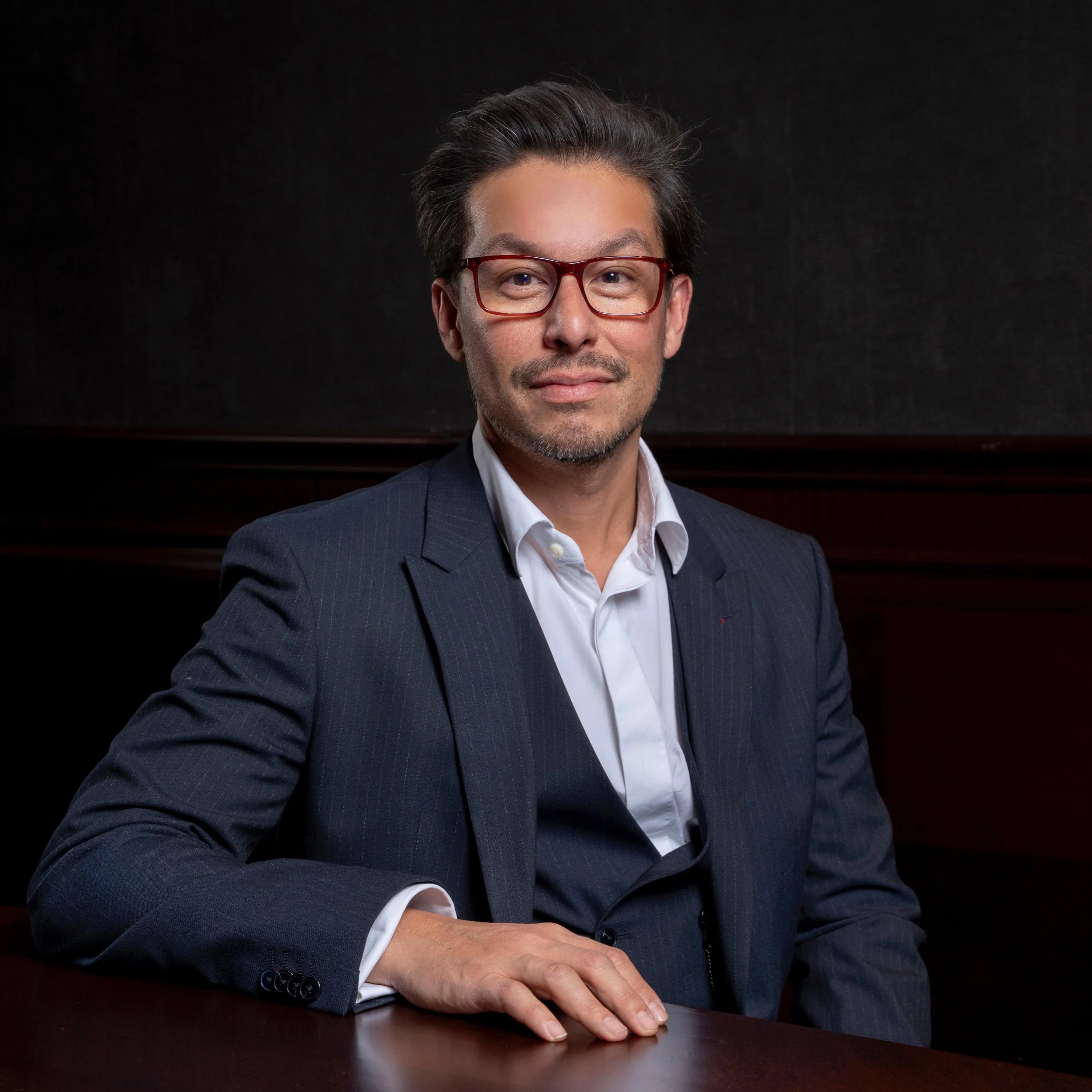 A man wearing glasses in a dark suit with a white shirt sitting at a table with his hand resting on the surface, against a dark background.