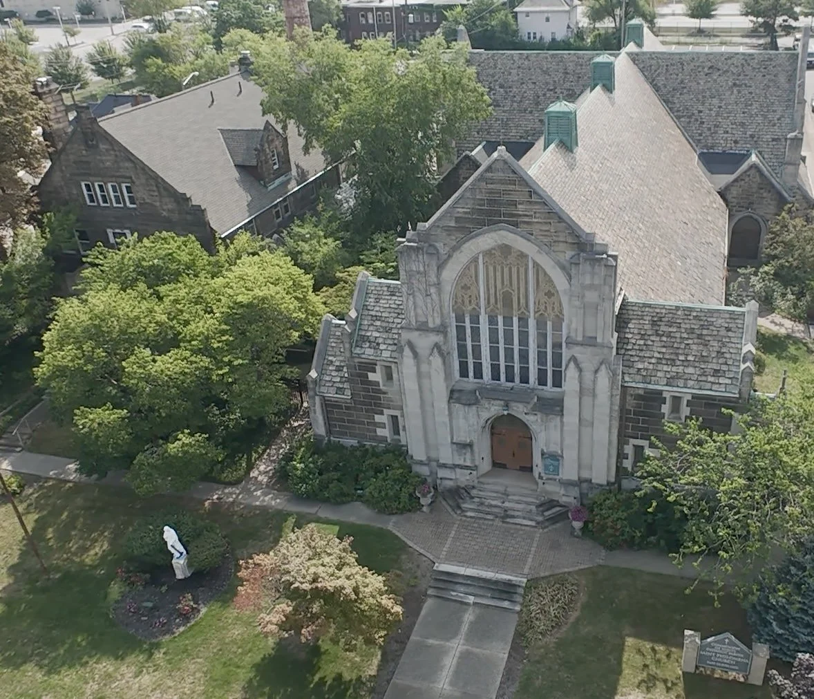An aerial view of a church with a large stained glass window and stone architecture, surrounded by trees and residential houses.