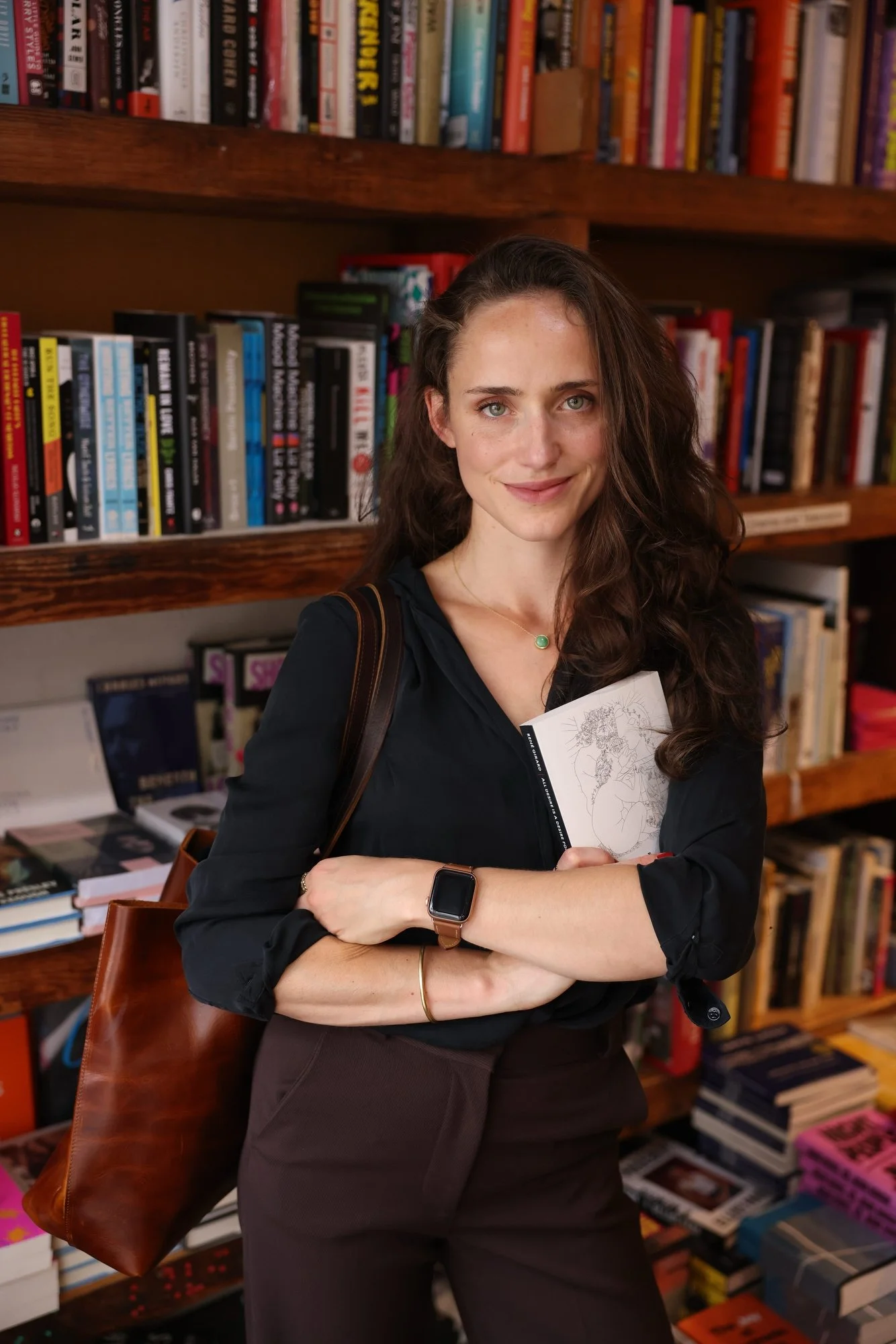 A woman with long brown hair and a black shirt standing in front of bookshelves filled with books, holding a book with a line drawing on the cover, wearing a smartwatch and a green pendant necklace.