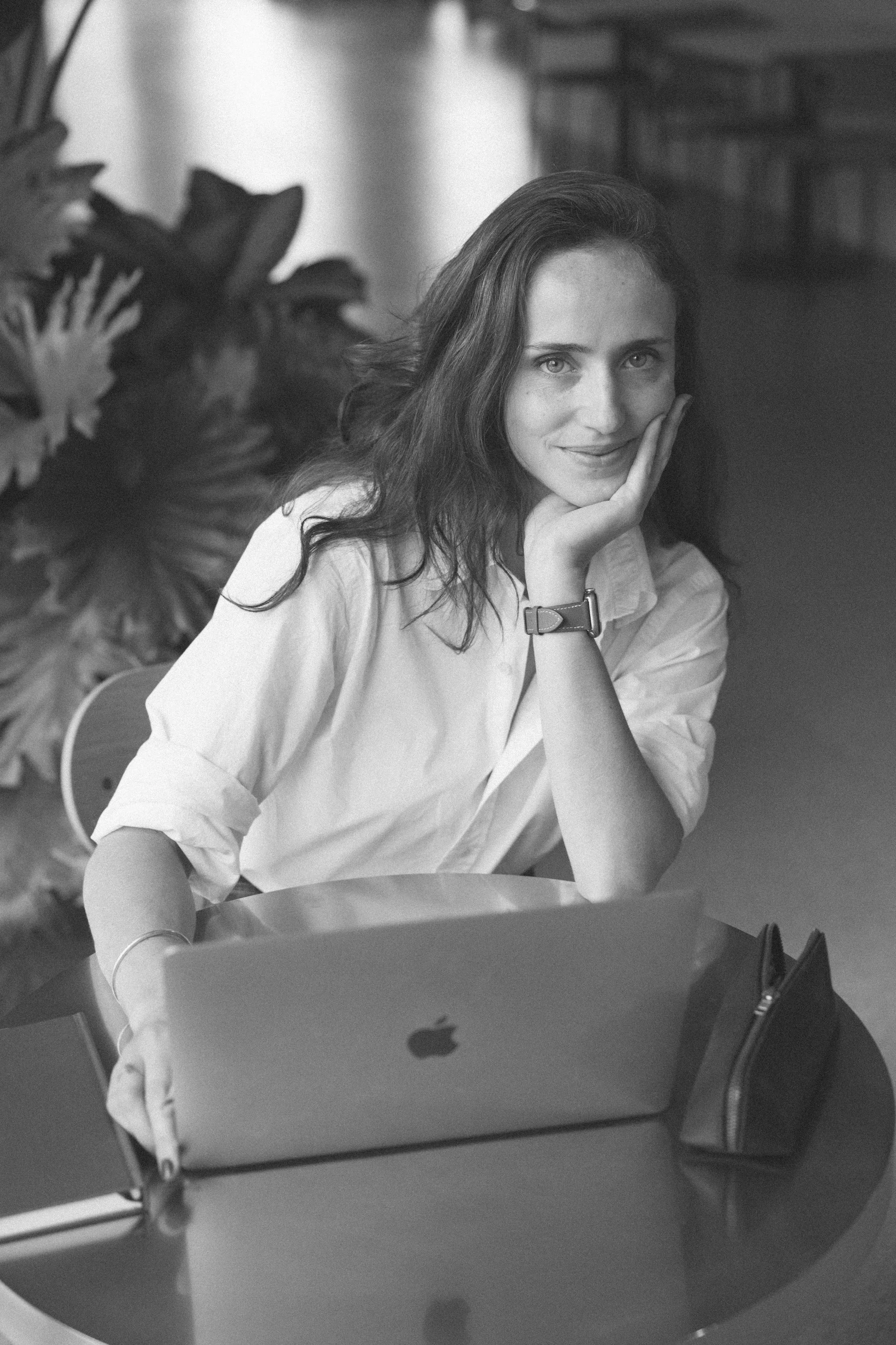 A woman with long, wavy hair sits at a table with an open laptop in front of her, smiling gently with her face resting on her hand, in a black and white photo.