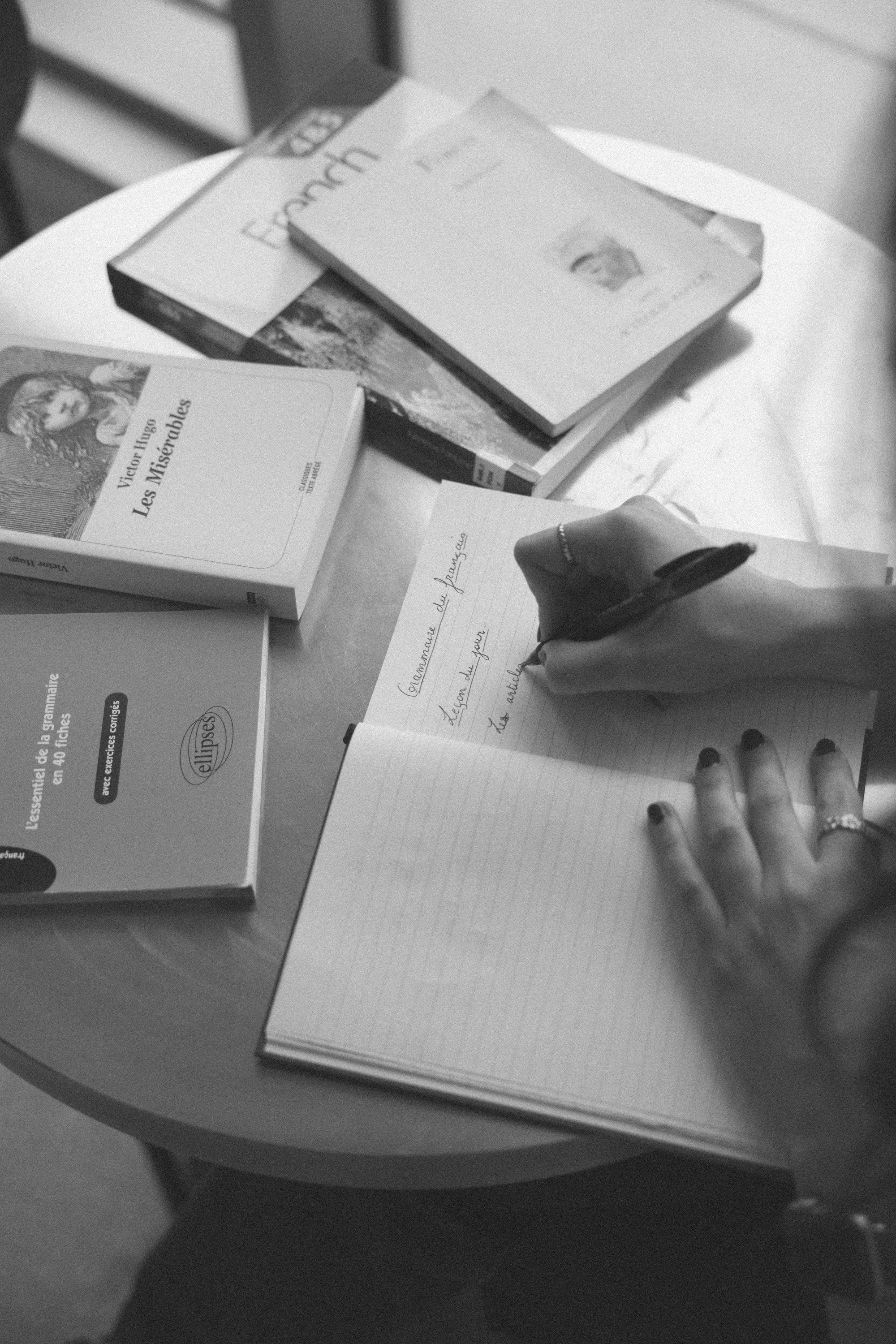 A person taking notes in a notebook on a round table with books about French language and literature, including Victor Hugo's Les Misérables, and a booklet titled 'L'essentiel de la grammaire'.