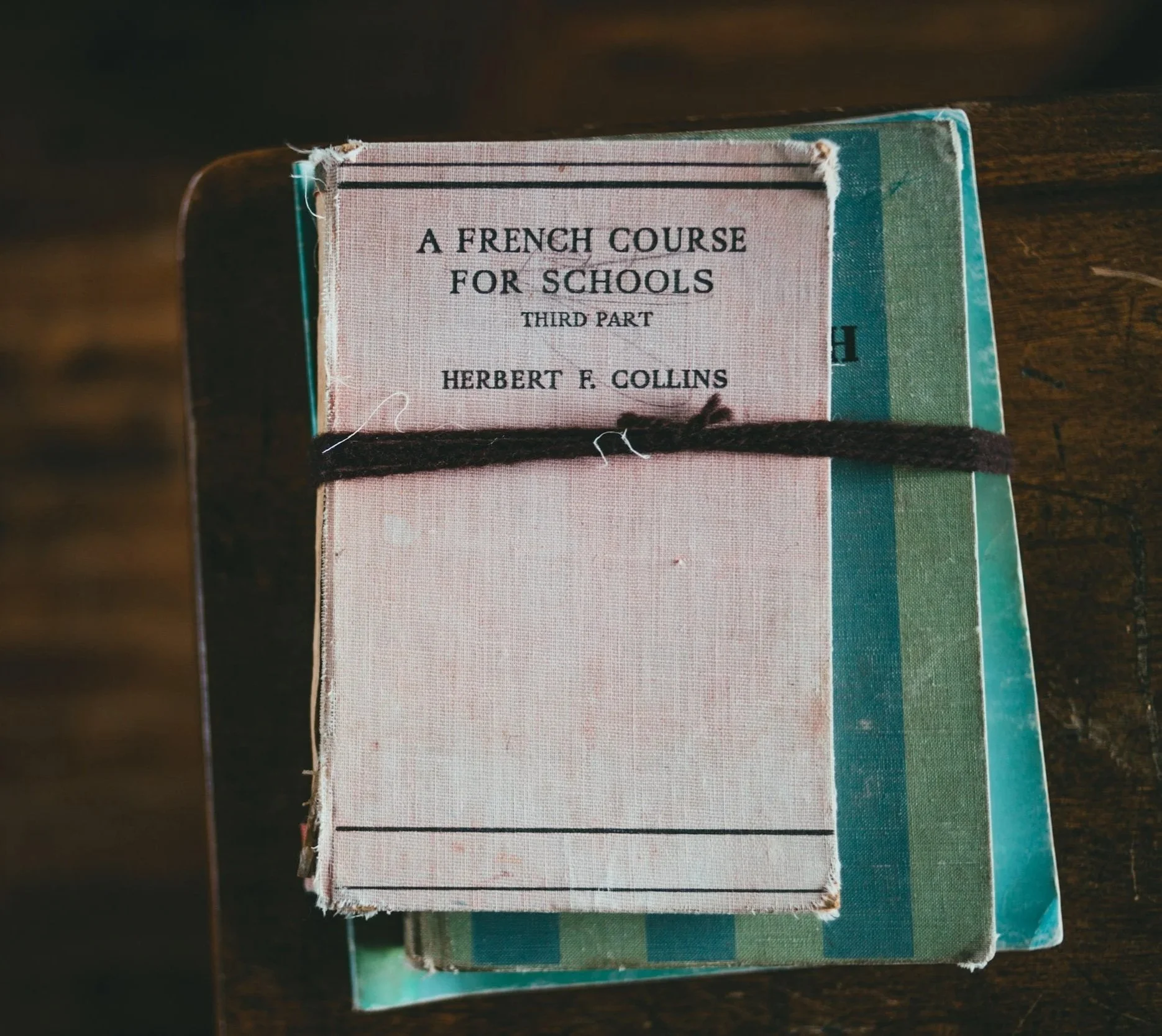 A stack of worn books with the top book titled 'A French Course for Schools, Third Part, Herbert F. Collins.' The books are tied together with a dark string, and they are placed on a wooden surface.