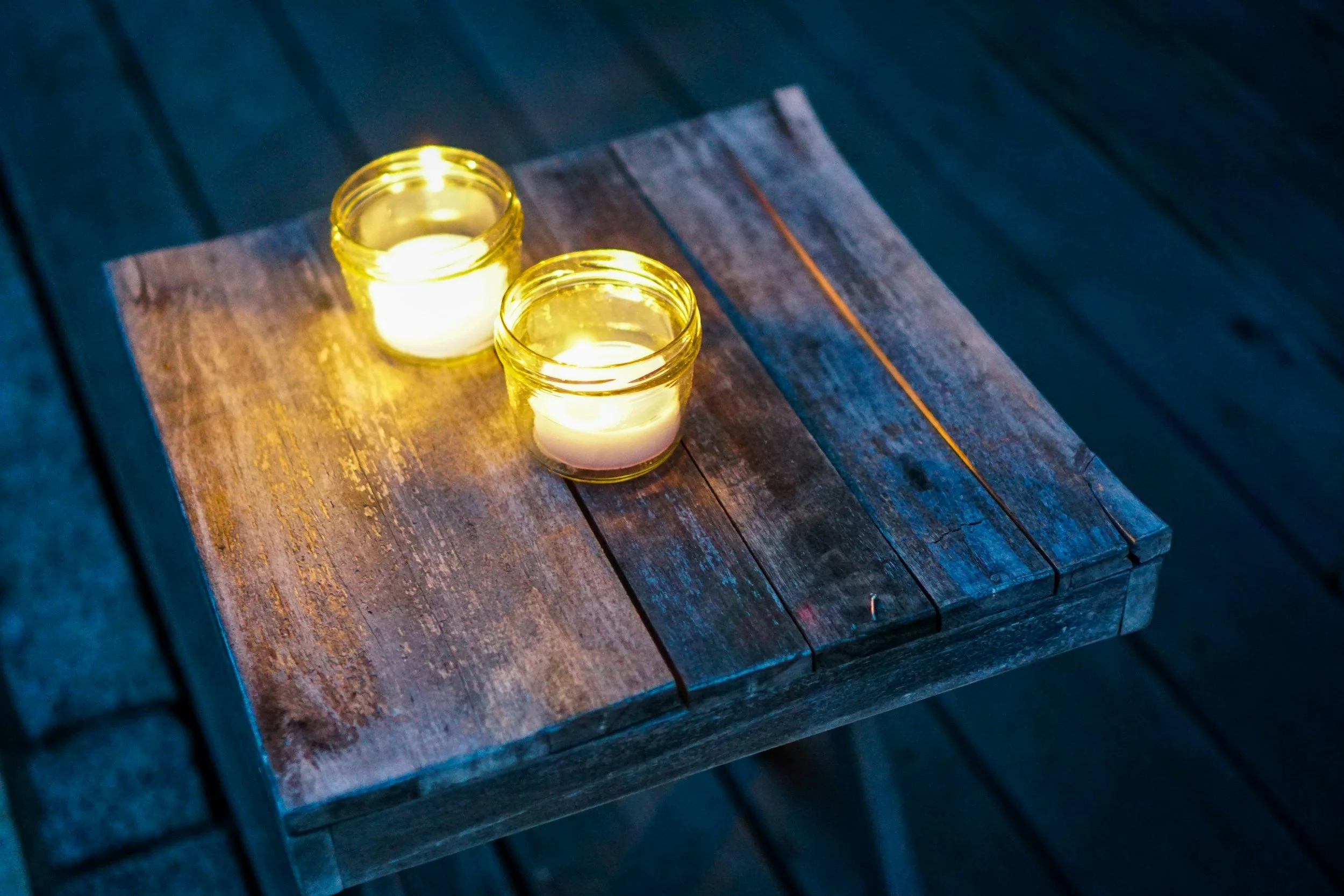 Two lit candles in glass holders on a weathered wooden table at night.