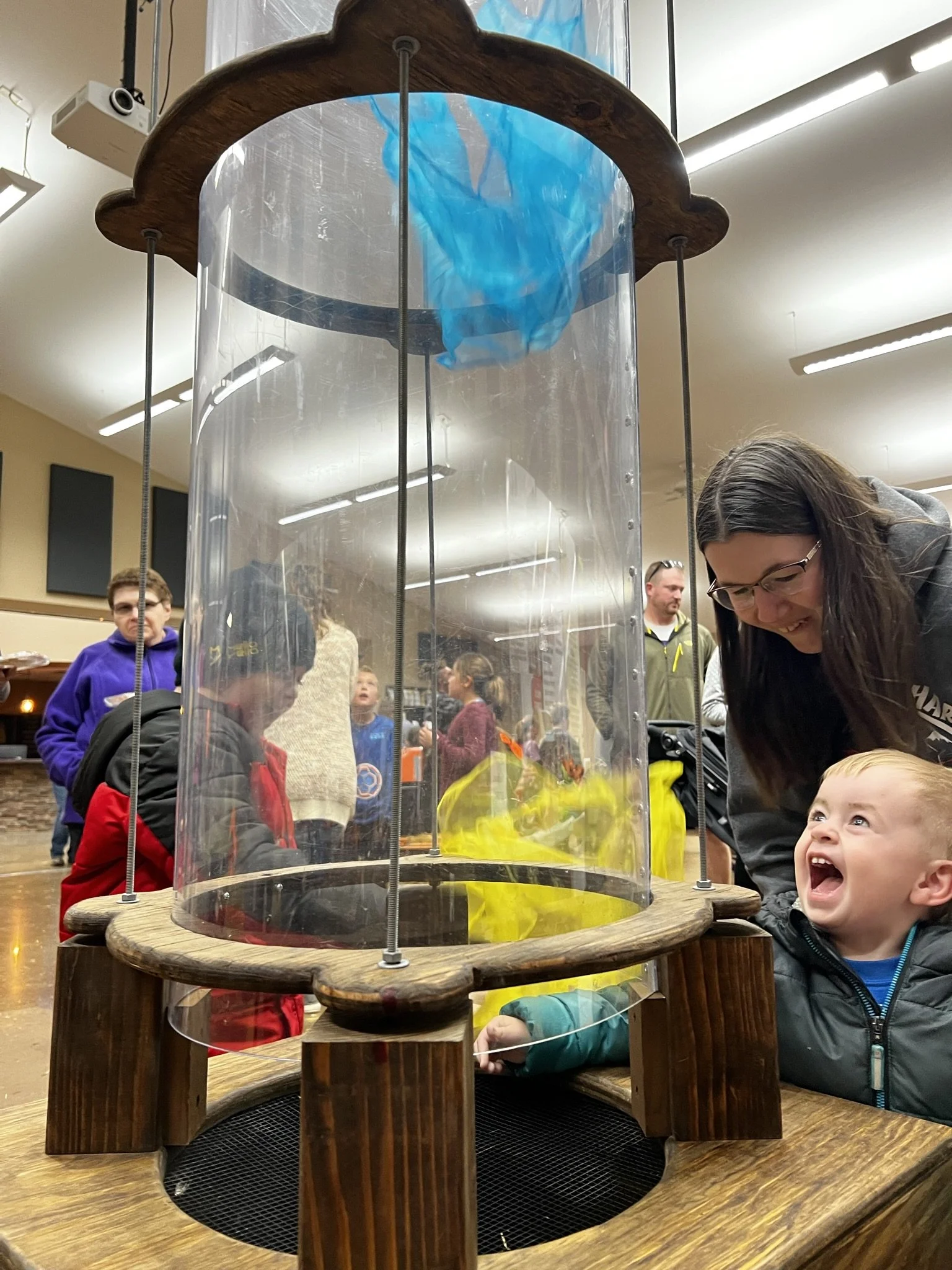 A group of children and adults gathered around a large tabletop science experiment involving a bottle with a blue and yellow liquid inside, enclosed in a transparent cylindrical shield with a wooden frame, in a well-lit indoor space.