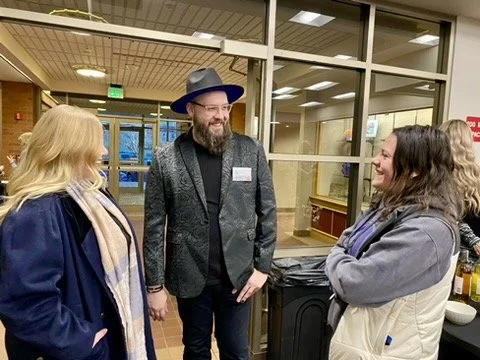 Three people talking and smiling in an indoor setting with glass windows and a trash can in the background.