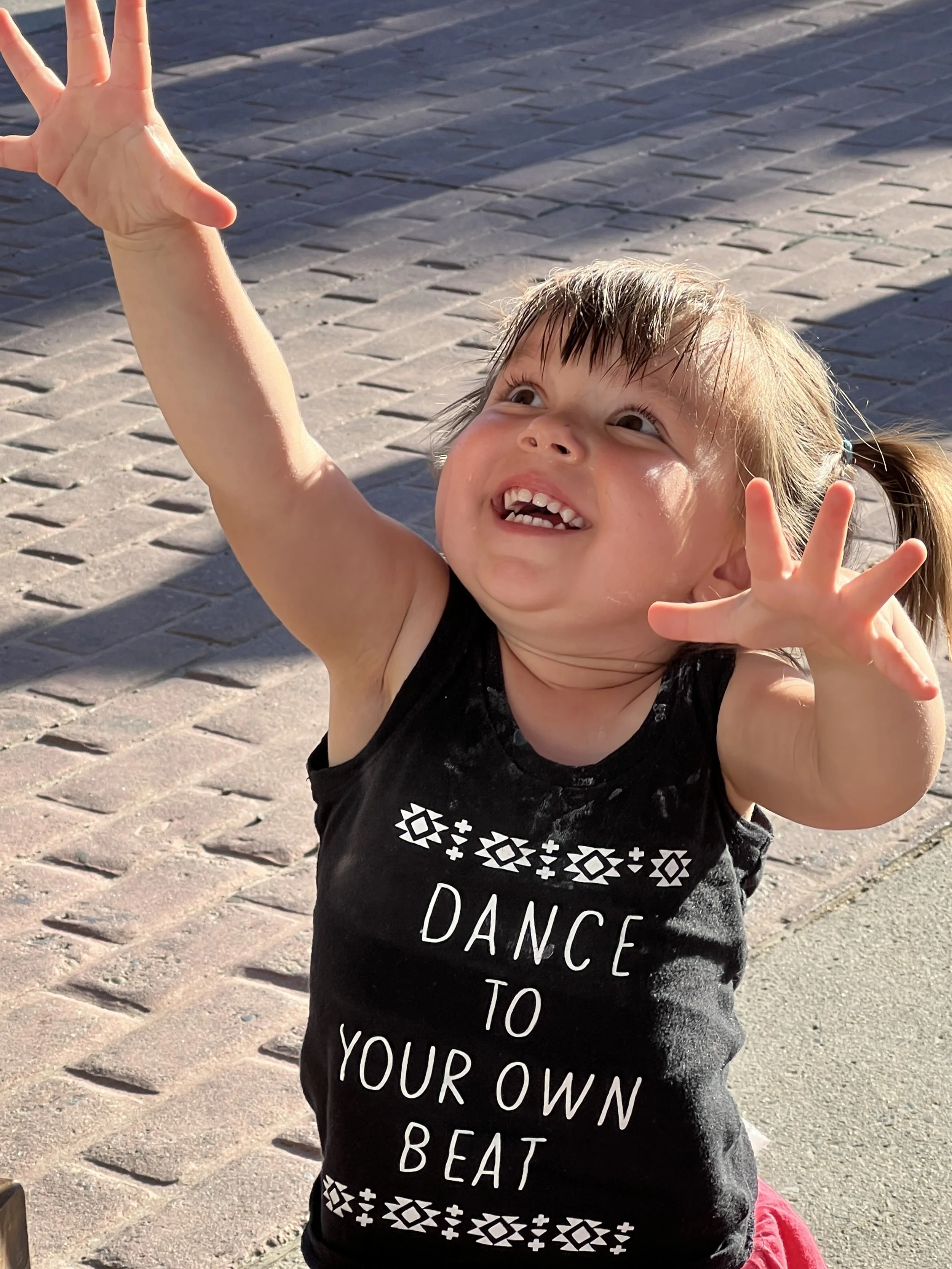 A young girl with light brown hair in pigtails and a big smile, reaching upward with both hands, outdoors on a sunny day with a brick sidewalk in the background, wearing a black sleeveless shirt with the text 'Dance to Your Own Beat' and a red skirt.