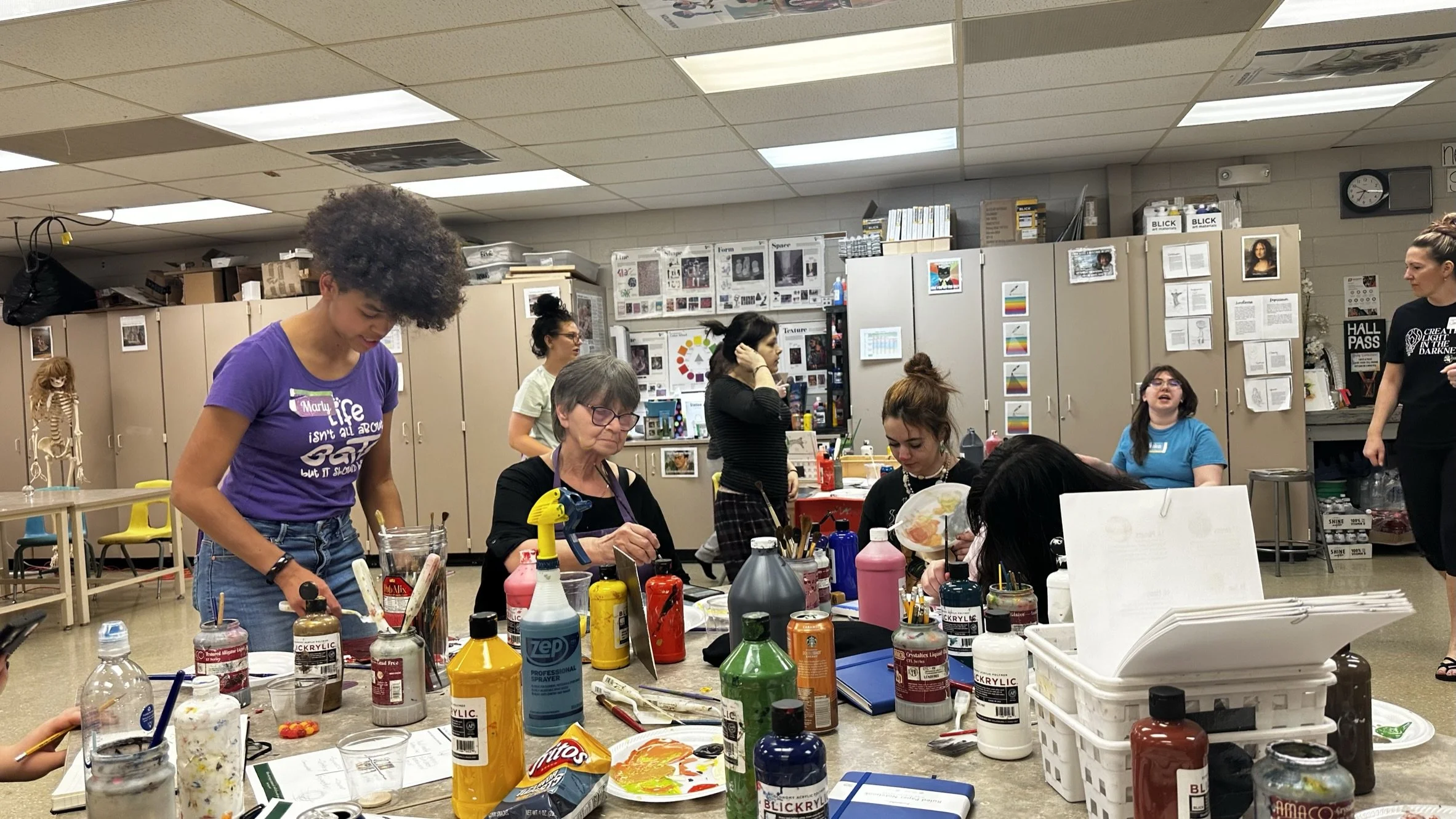 People participating in an art or craft class in a classroom or studio. Multiple individuals are engaged in painting or other creative activities at tables filled with art supplies, including bottles of paint, brushes, and palettes. The background shows cabinets, posters, and art-related information on the walls.