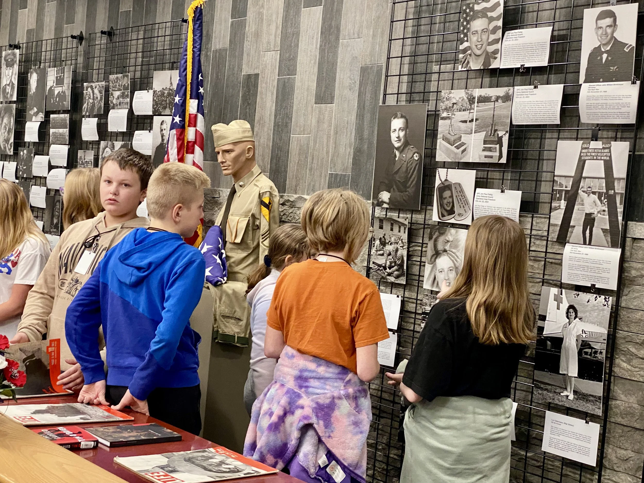 The image shows a group of children gathered at a memorial or museum display with black-and-white photographs and descriptions of military personnel, including a mannequin dressed in a military uniform and an American flag.