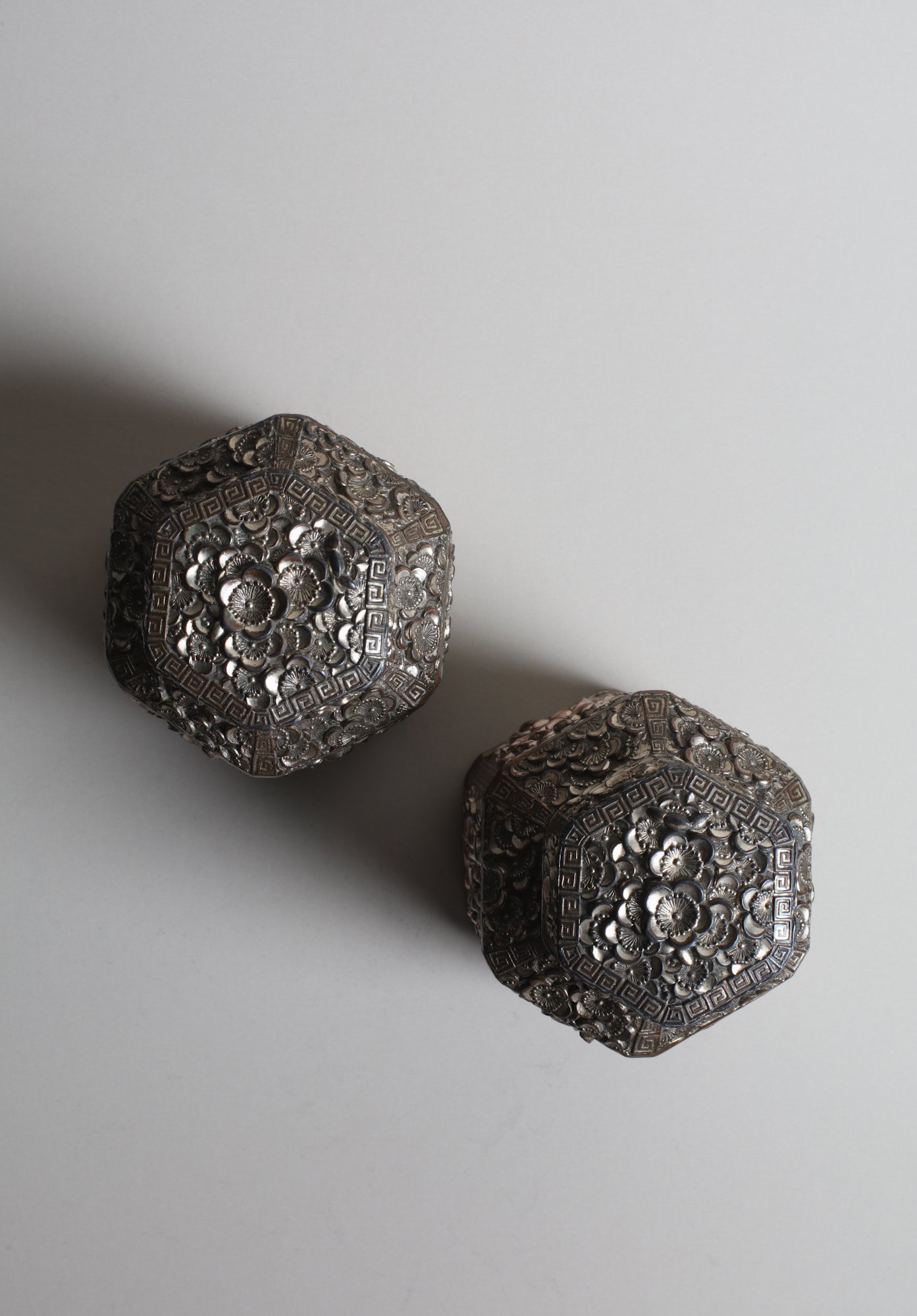 Close-up of two ornate repousse, silver-colored metal chinese tea caddy lids with intricate floral and geometric patterns, arranged on a plain, light gray background.