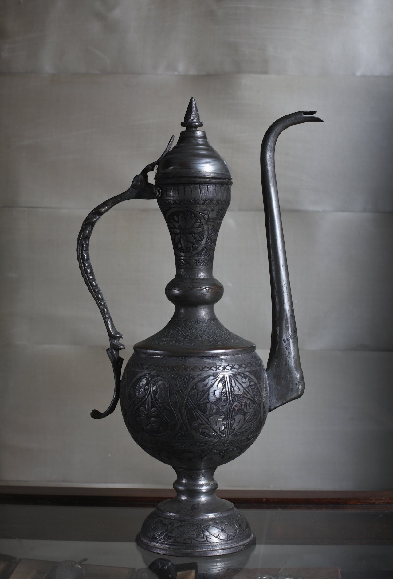 A tall, ornate metal teapot with detailed patterns and a long, curved spout, displayed against a plain background.