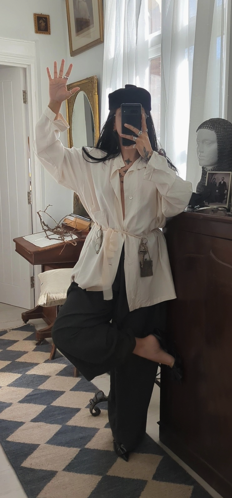 A woman taking a mirror photo in a antique-style room, wearing a black hat, cream oversized shirt with a belt, black pants, and black shoes. The room has framed art on the wall, a mirror, a wooden table with decor, and a mannequin head with hair.