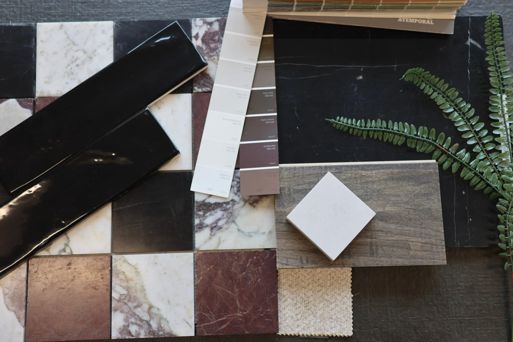 Modern European Luxury interior design flat lay, featuring marble mosaic and wood tile samples, a paint color swatch, a small square tile, and some green fern leaves on a dark background.