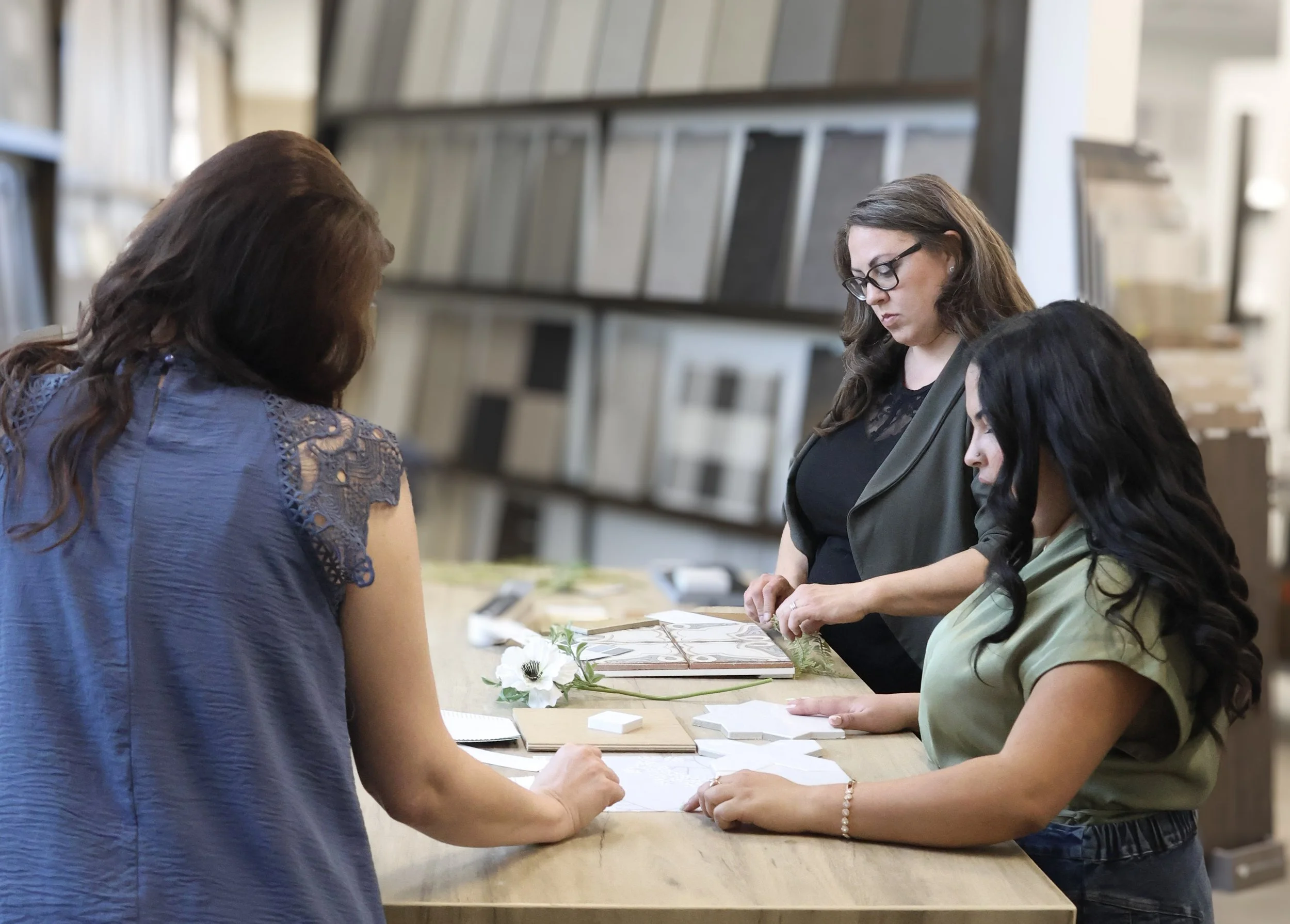 Three women are standing at a counter, looking at design samples or swatches, in what appears to be a furniture or home decor store.