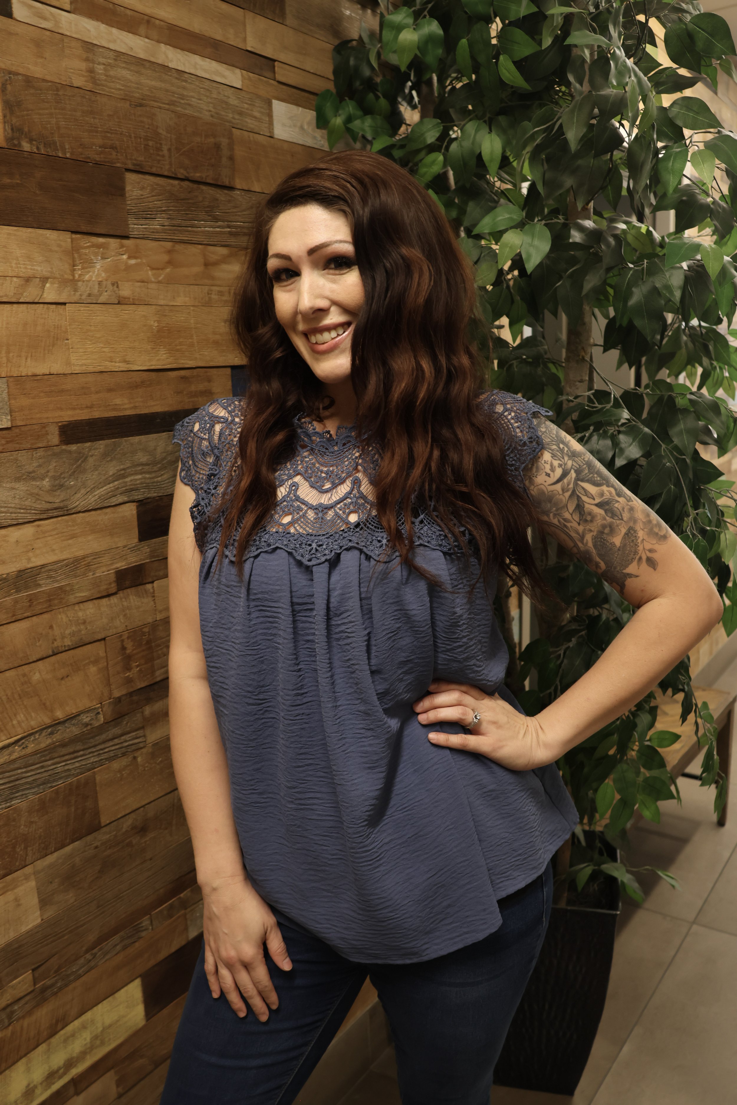 A woman with long wavy brown hair, wearing a blue lace top and dark jeans, stands indoors beside a wooden wall and a potted plant. She is smiling and posing with her right hand on her hip.