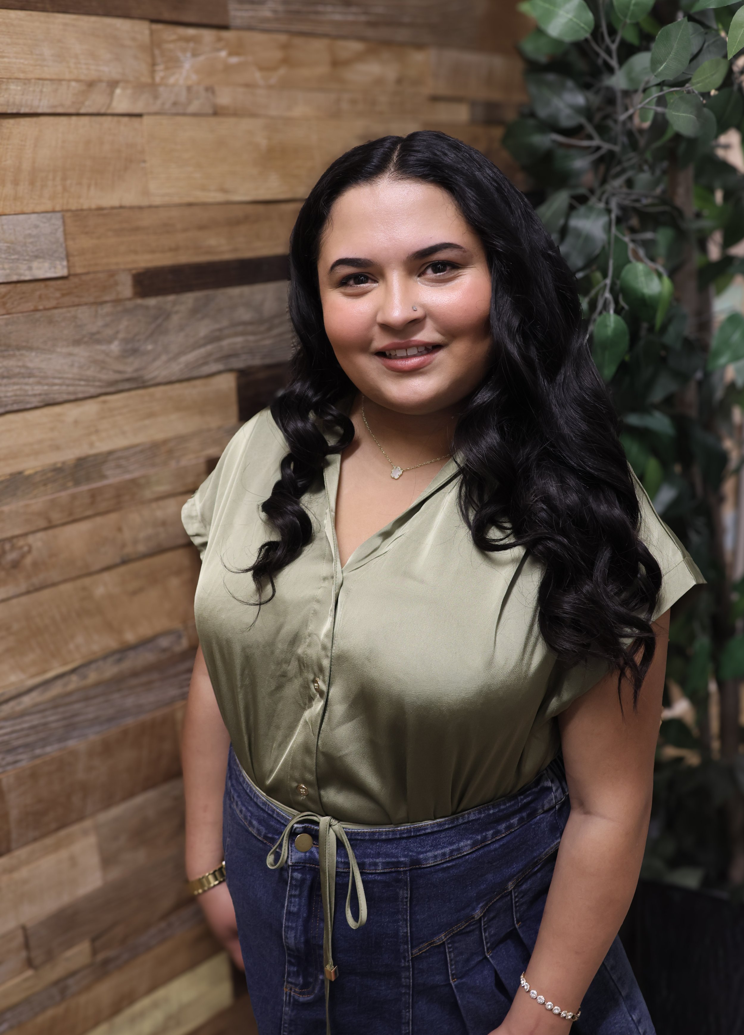 A woman with long black curly hair, wearing a light green blouse with a tied waist, dark blue jean skirt, and jewelry, standing in front of a wooden wall and green plants.