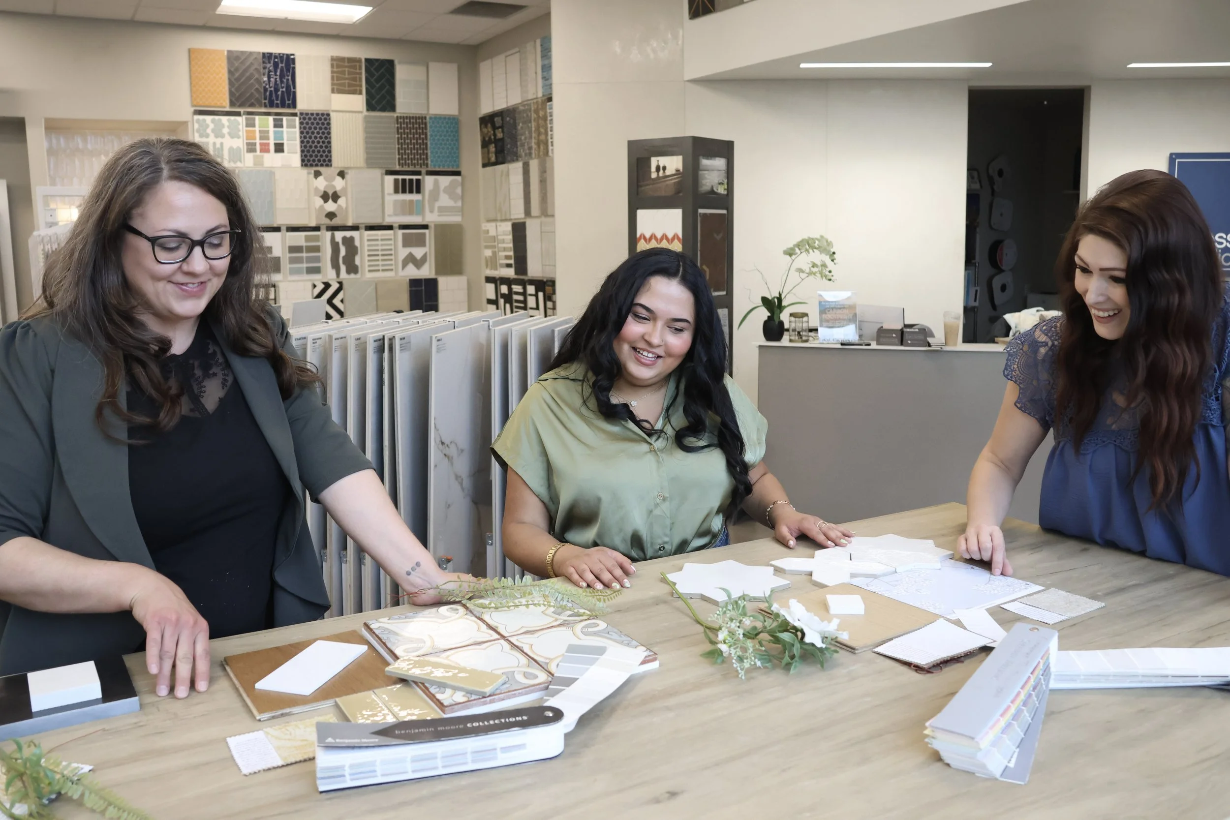 Three women are sitting at a table in a showroom, looking at and discussing various sample tiles, paint chips, and color palettes for interior design or decorating projects. The background includes wall samples and design displays.