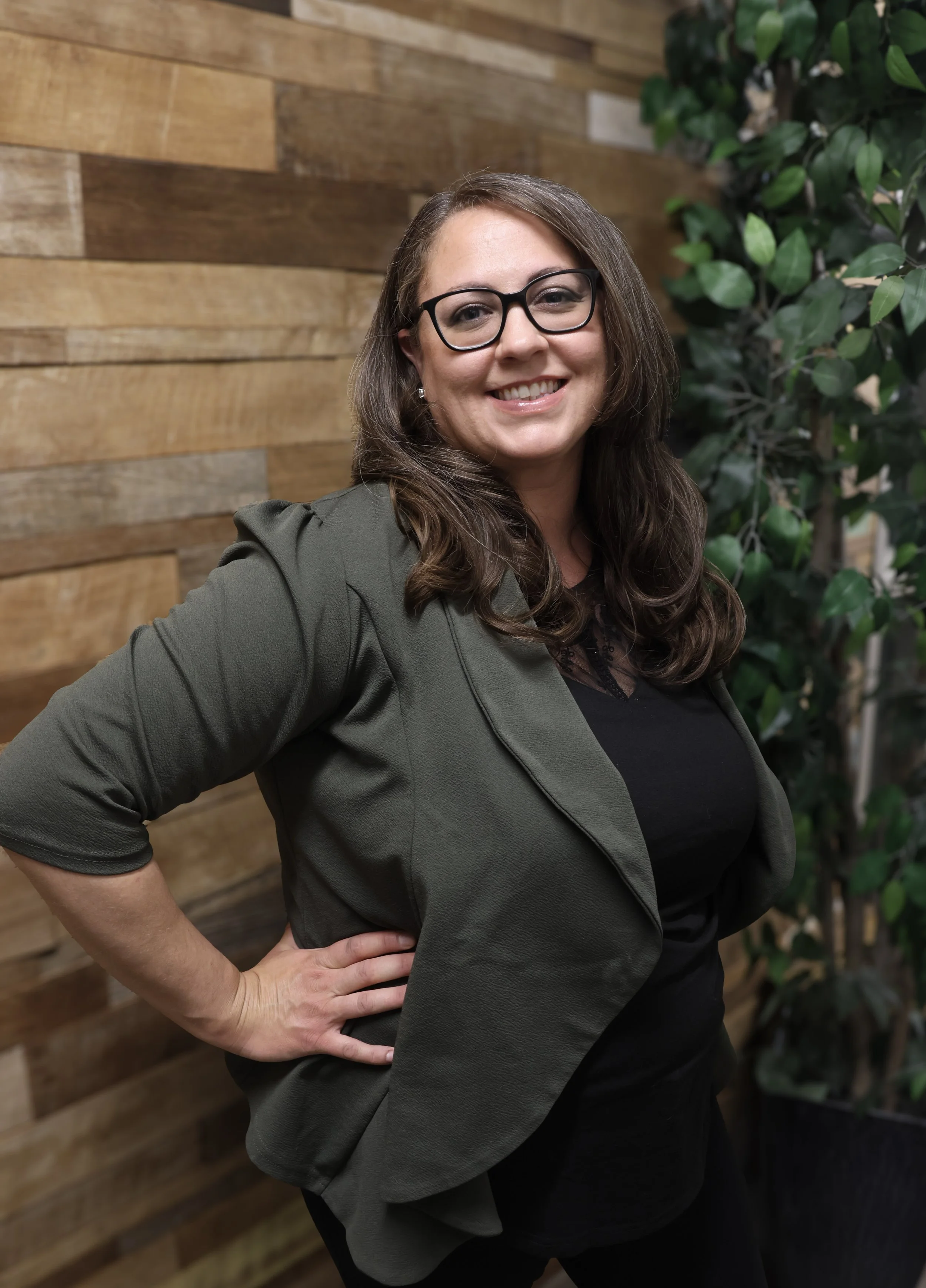 A woman with long brown hair wearing glasses, a dark lace top, and a dark jacket standing with her right hand on her hip in front of a wooden wall and a green leafy plant, smiling at the camera.