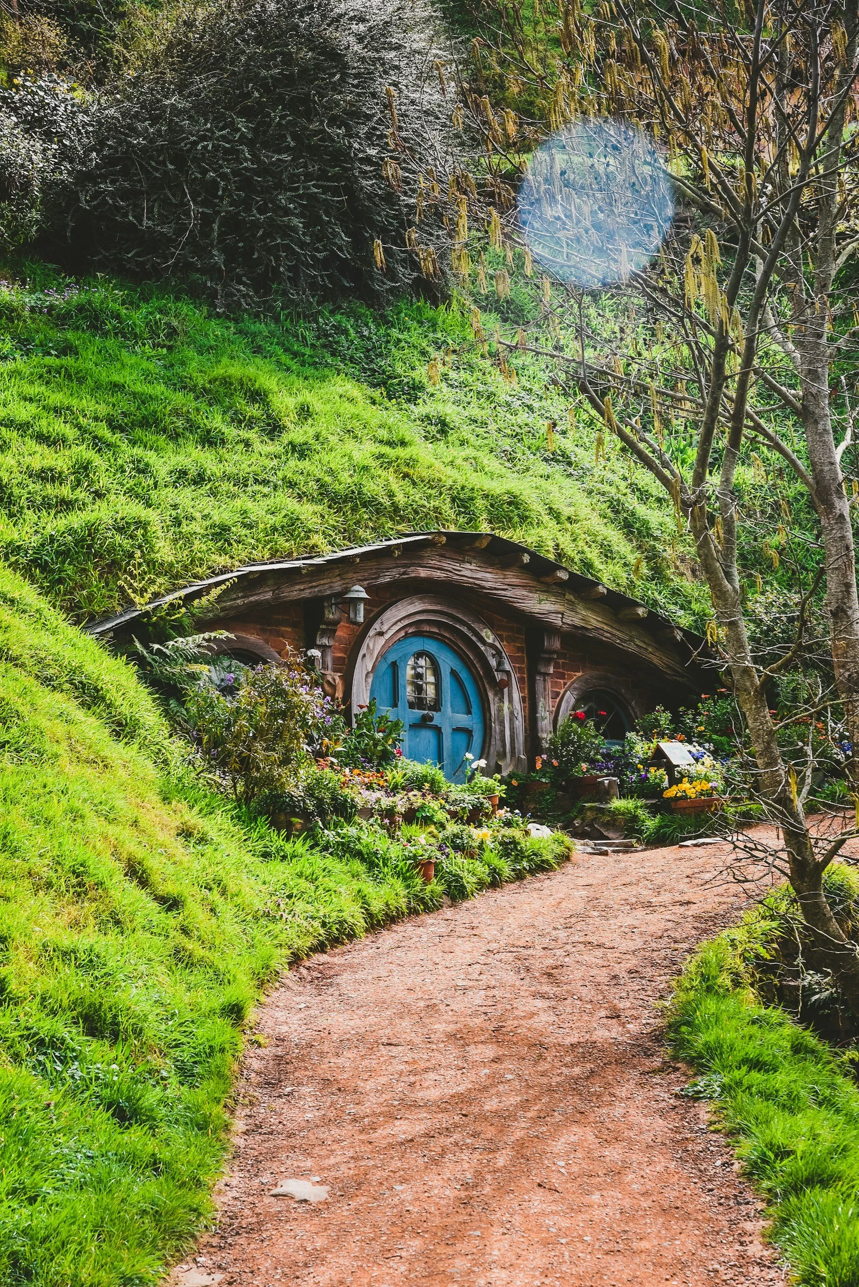 The image shows a hobbit house built into a lush, green hillside with a dirt path leading to a blue door, surrounded by flowers and plants.