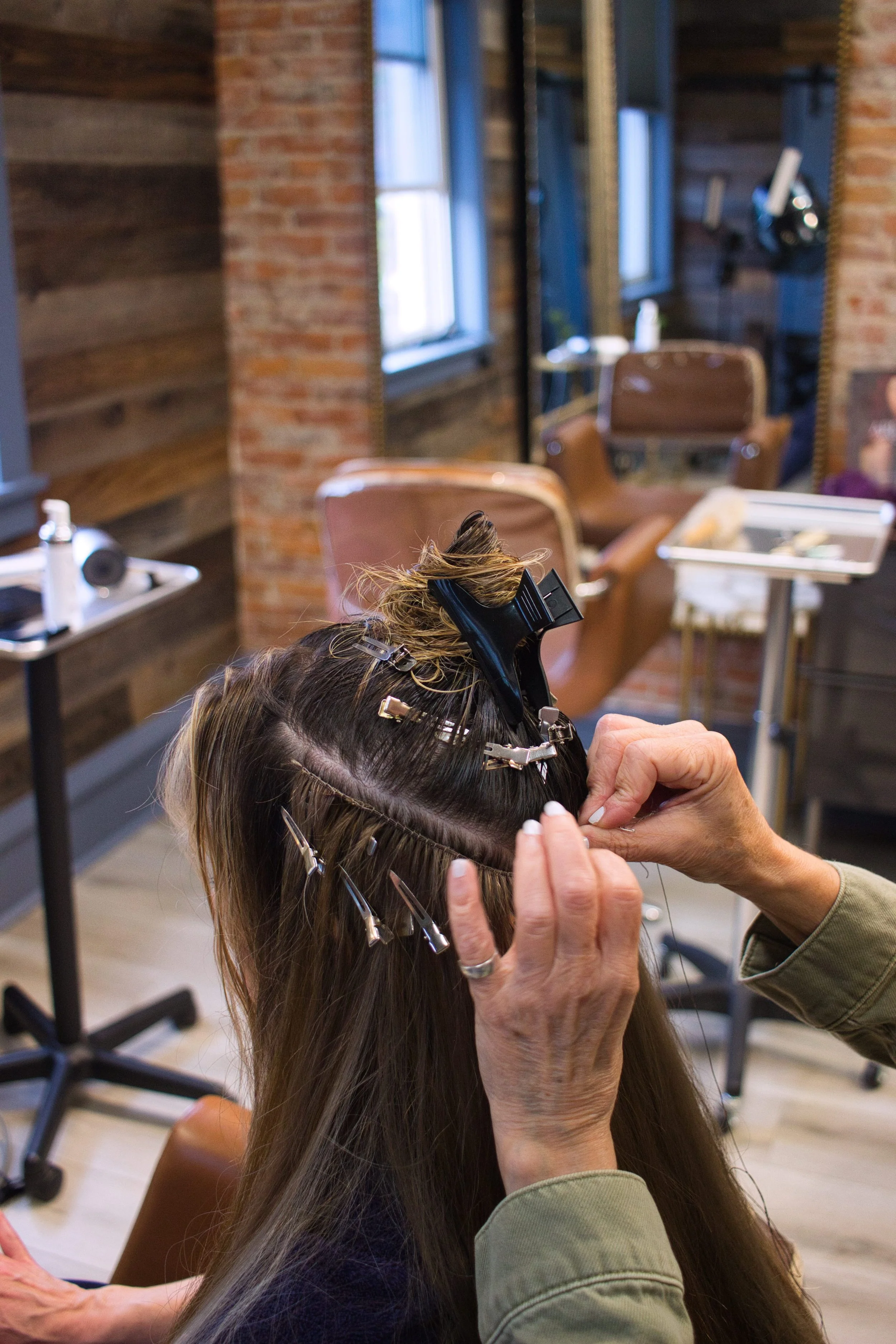A hairstylist is applying hair extensions or preparing hair with clips in a salon with brown chairs, wooden walls, and large windows.