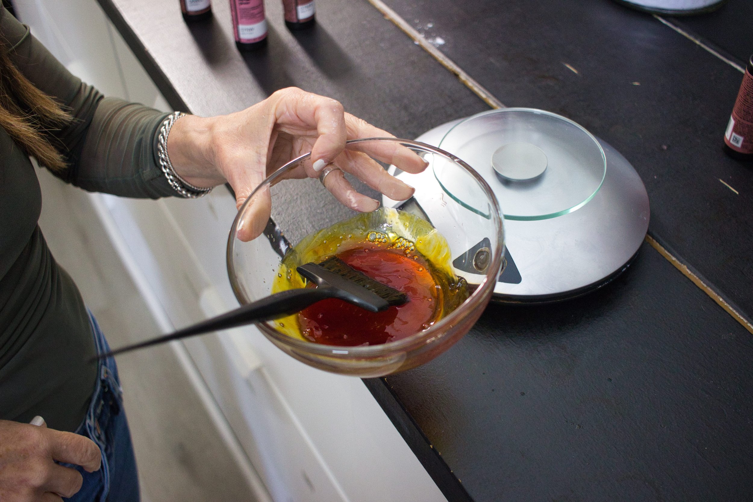 A person holding a clear glass mixing bowl with yellow and red liquids inside, with soap bubbles on the surface, in a kitchen or food prep area. The person is wearing a silver bracelet and has painted nails.
